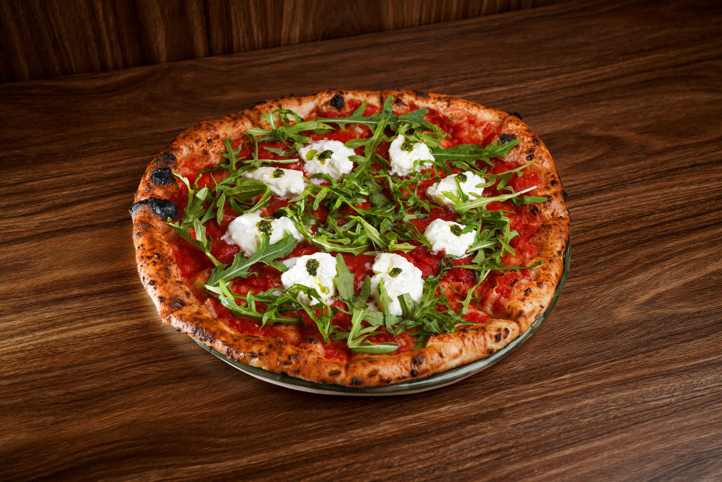 A freshly baked pizza on a wooden table, topped with arugula, mozzarella, tomato sauce, and pesto drizzles.
