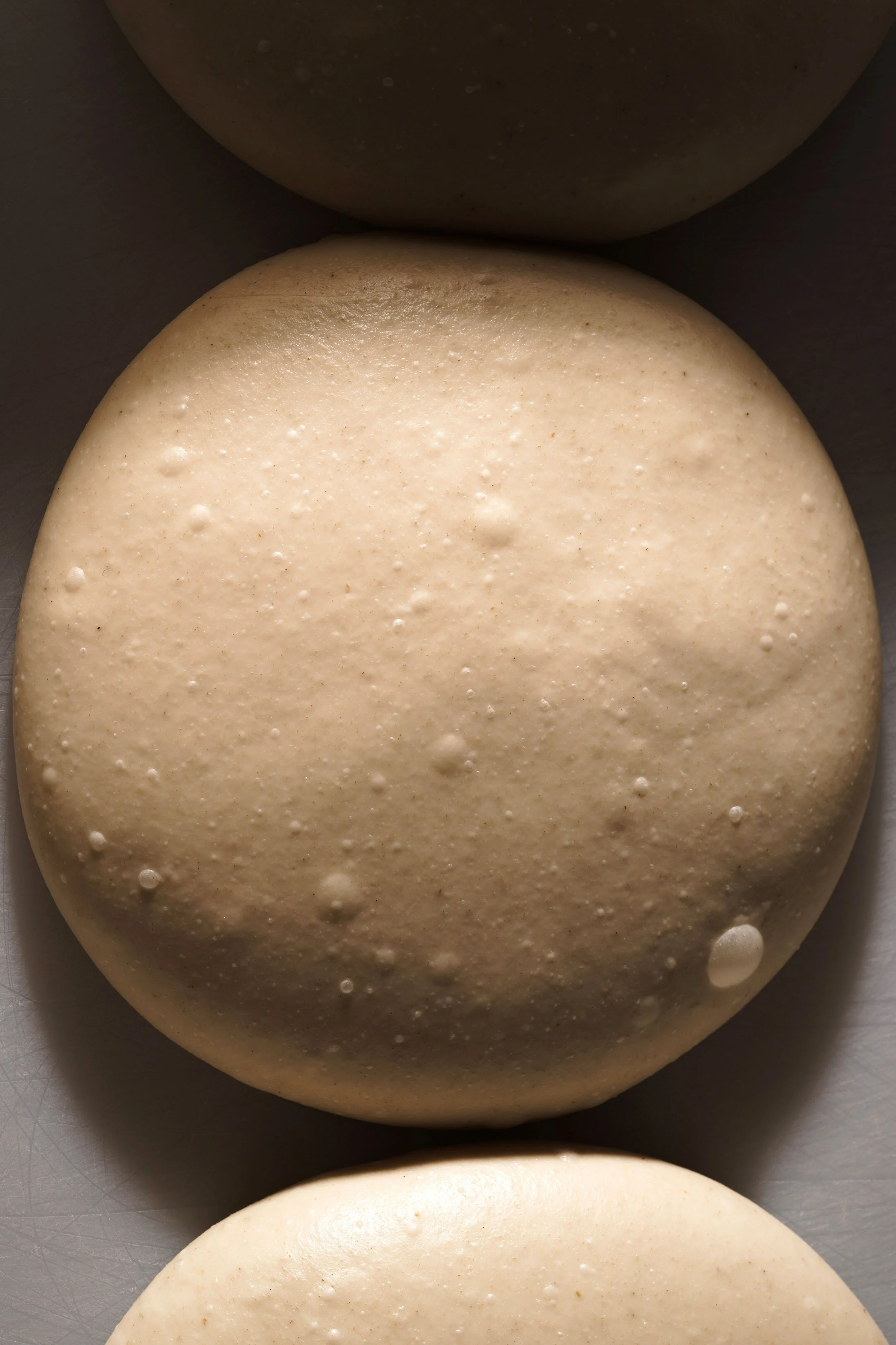 Close-up photo of several round, smooth dough balls with a slightly textured surface, some with small bubbles, on a gray surface.