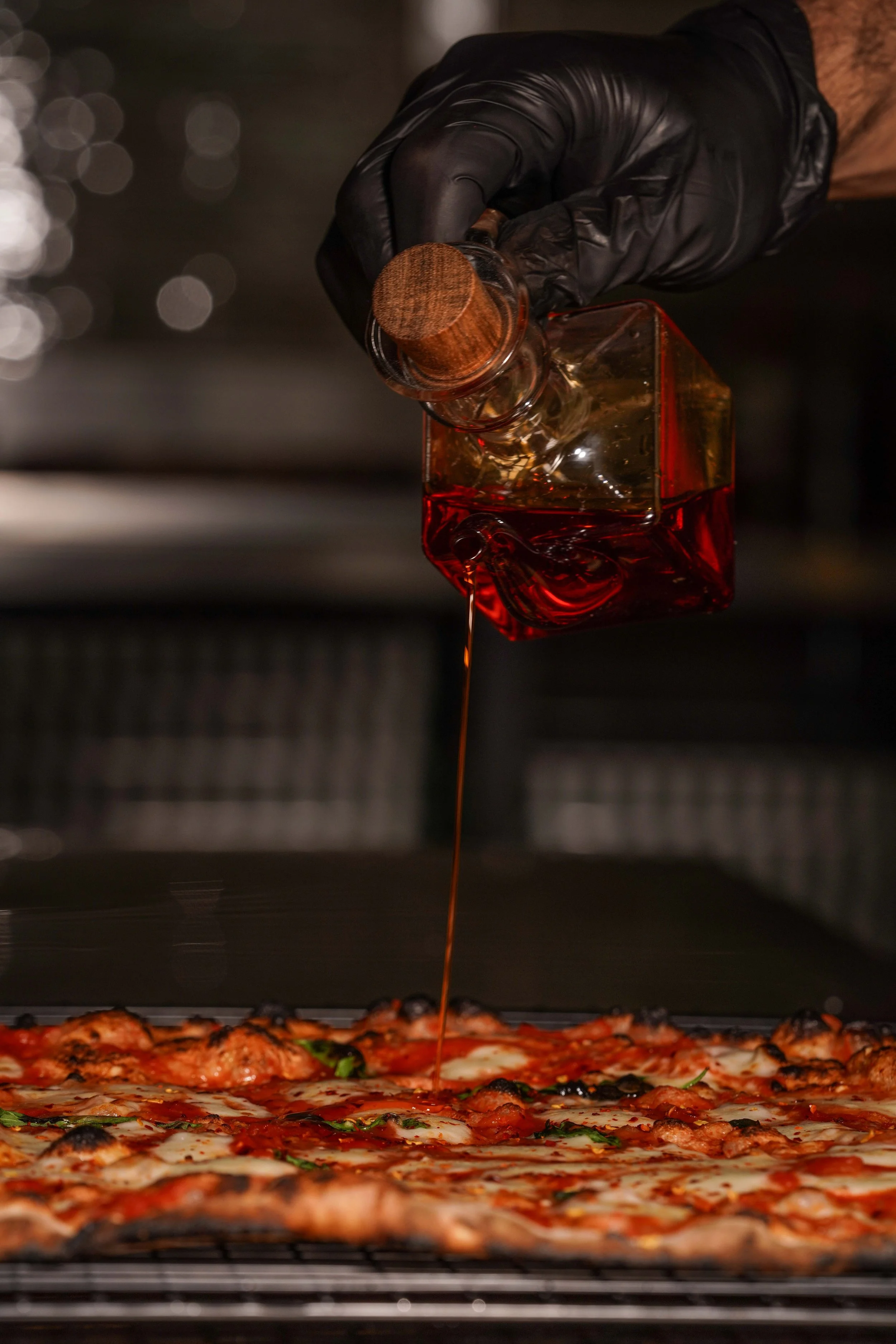 Close-up of a hand wearing a black glove pouring hot sauce on a pizza. The pizza has visible toppings including cheese, pepperoni, and green herbs. The background appears to be an oven with a blurred, bokeh effect.