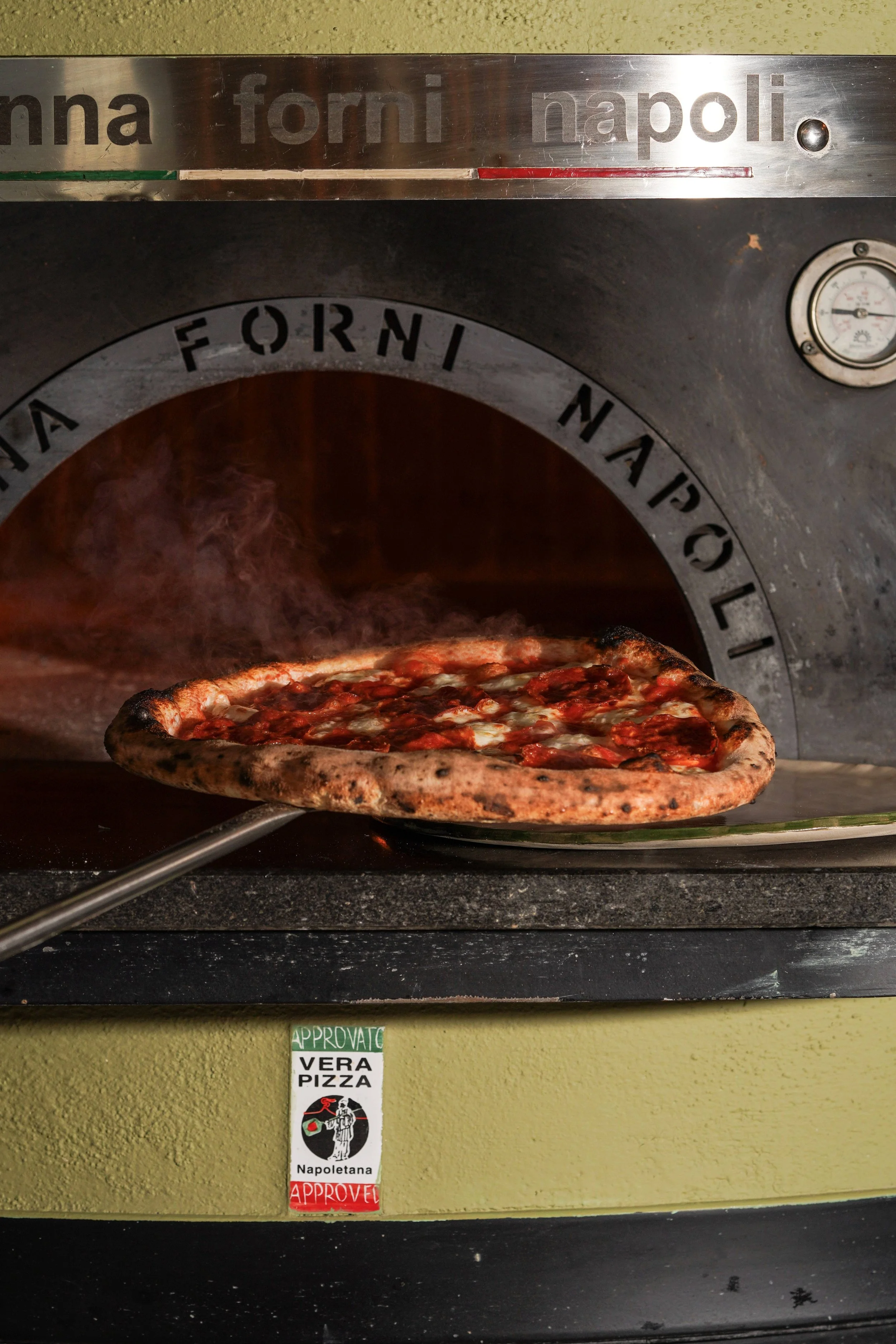 Pizza coming out of a pizza oven, with a sign below that says 'VERA PIZZA' and 'NAPOLETANA' with approval stickers.