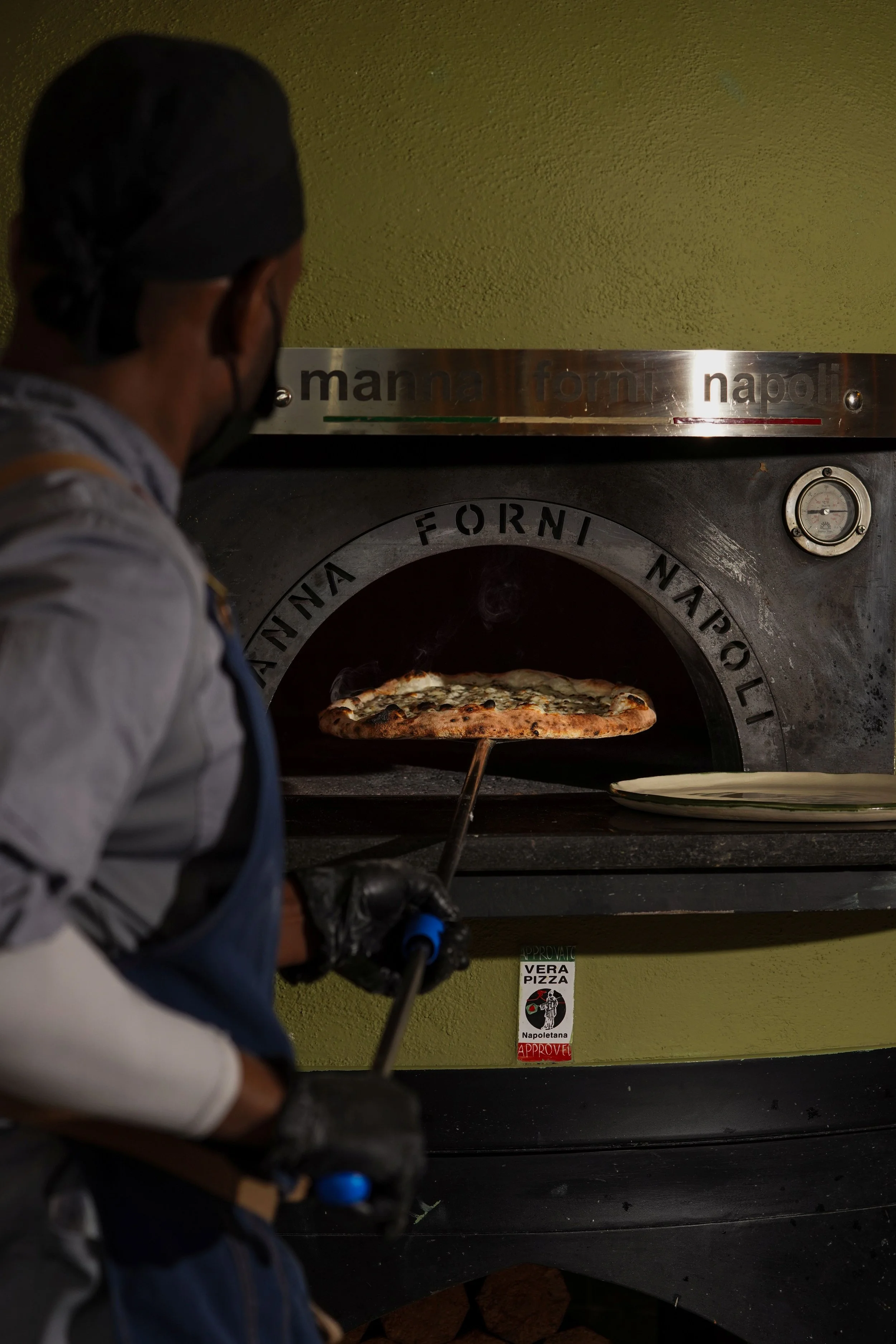 A person wearing a black hair covering, gray and blue clothing, and gloves, is opening a pizza oven with a pizza inside. The oven has individual Italian brand name and logo stickers, and a small temperature gauge on its front.