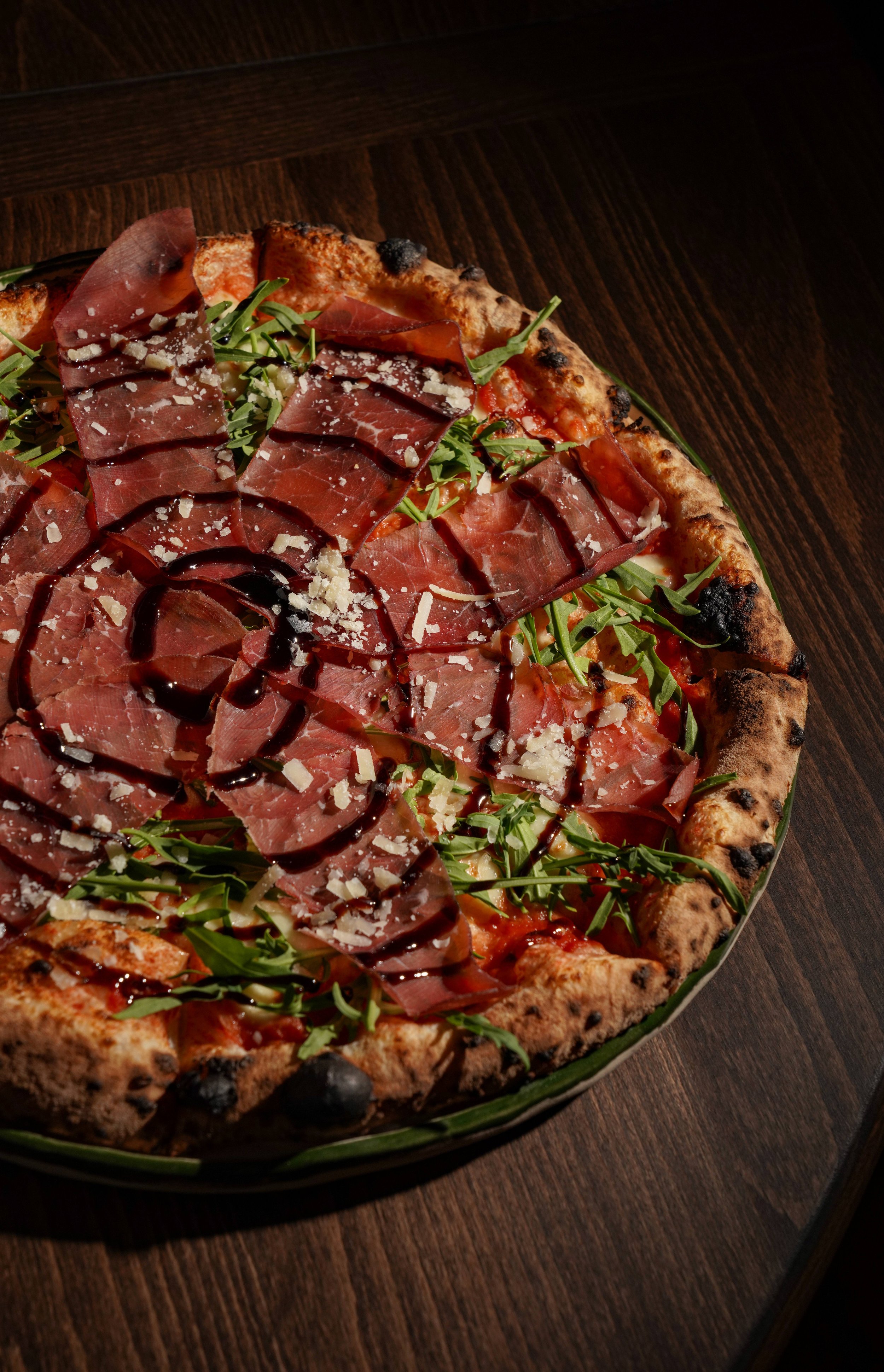 Close-up of a gourmet pizza topped with cured meats, arugula, shaved cheese, balsamic glaze, and a wood-fired crust on a wooden table