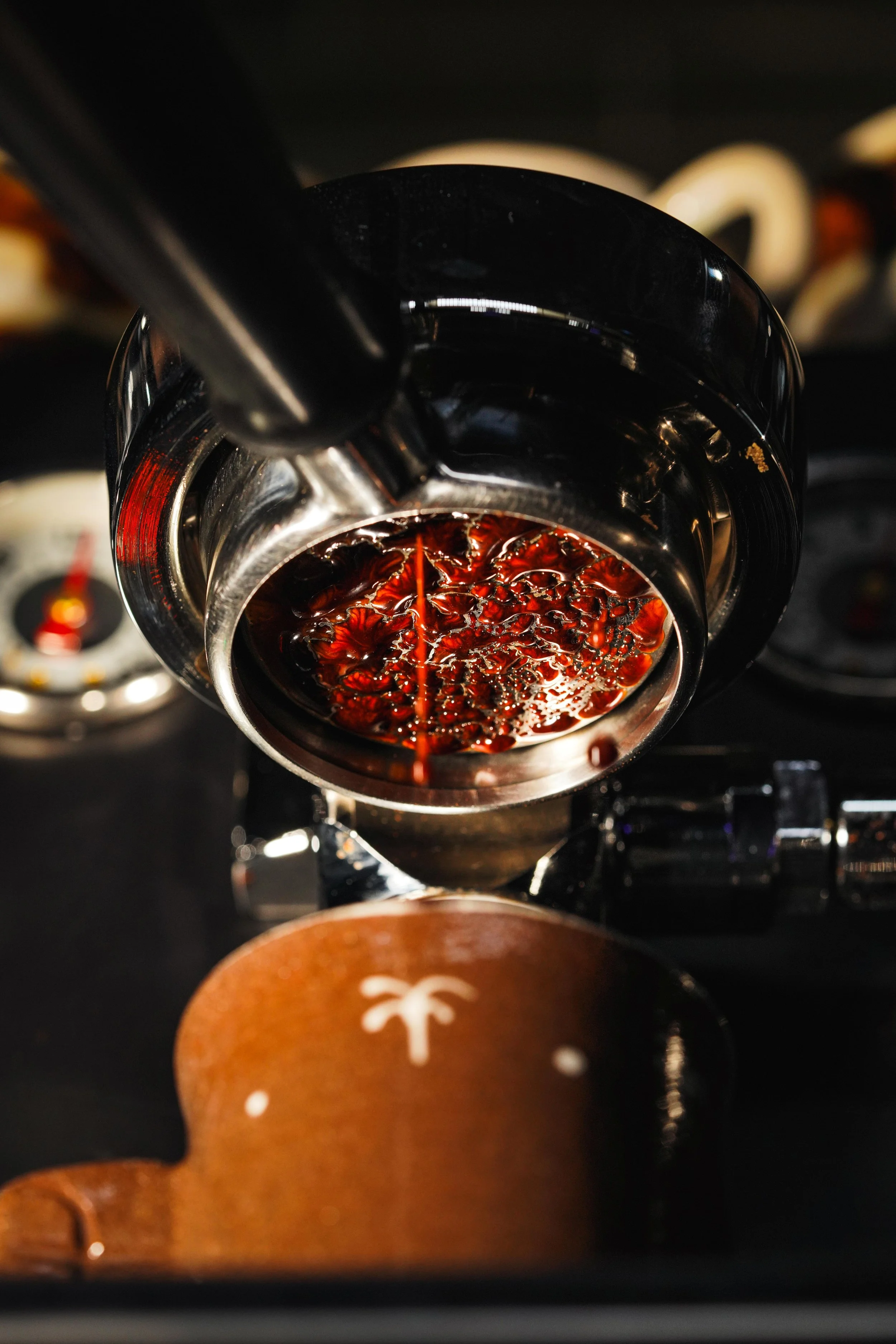 Close-up of a metallic espresso machine portafilter filled with brewed dark coffee, with a blurred background of coffee cups and accessories.