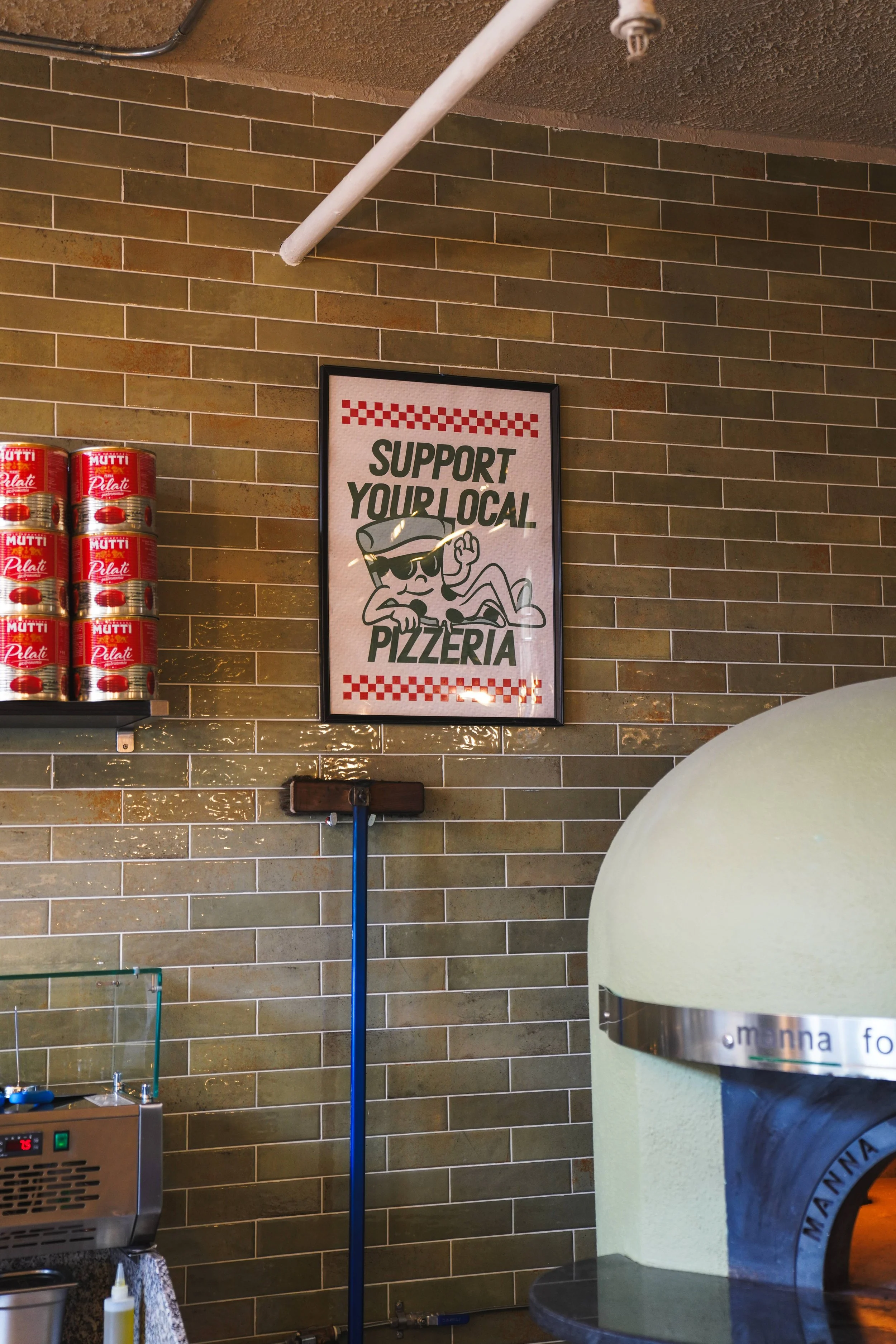 A pizza place interior featuring a framed sign that reads "Support Your Local Pizzeria" with a cartoon pizza mascot wearing sunglasses and holding a pizza. The wall behind includes a stack of canned tomatoes and a small appliance, possibly a pizza ov