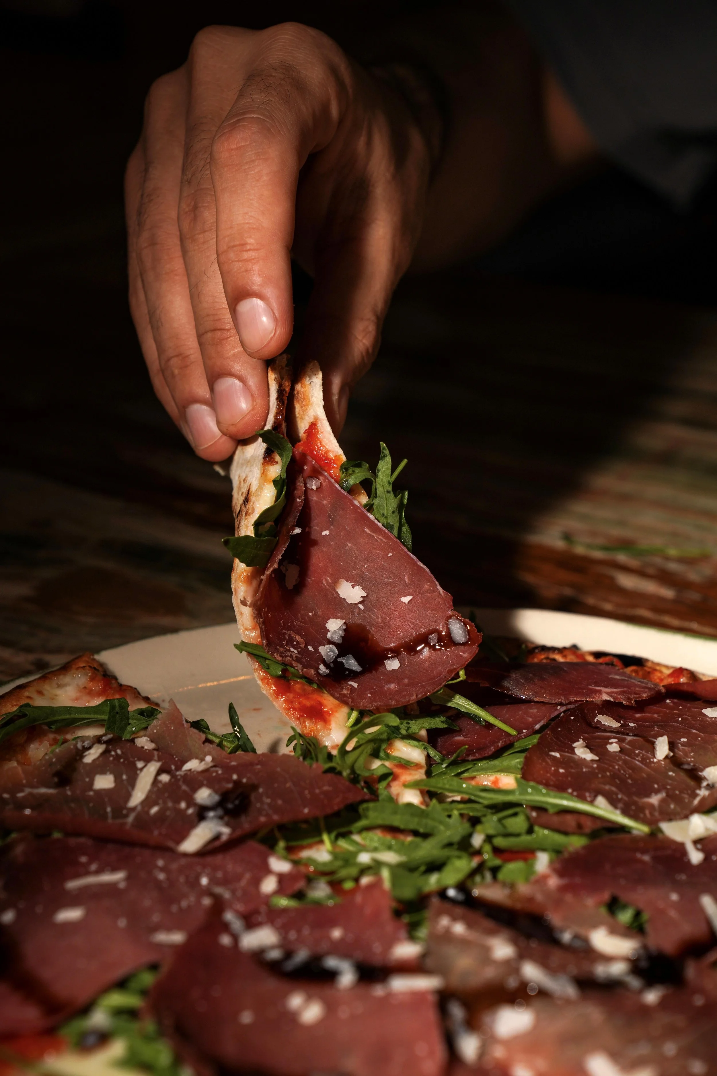 Close-up of a hand placing a slice of pizza topped with arugula, prosciutto, and grated cheese onto a plate.