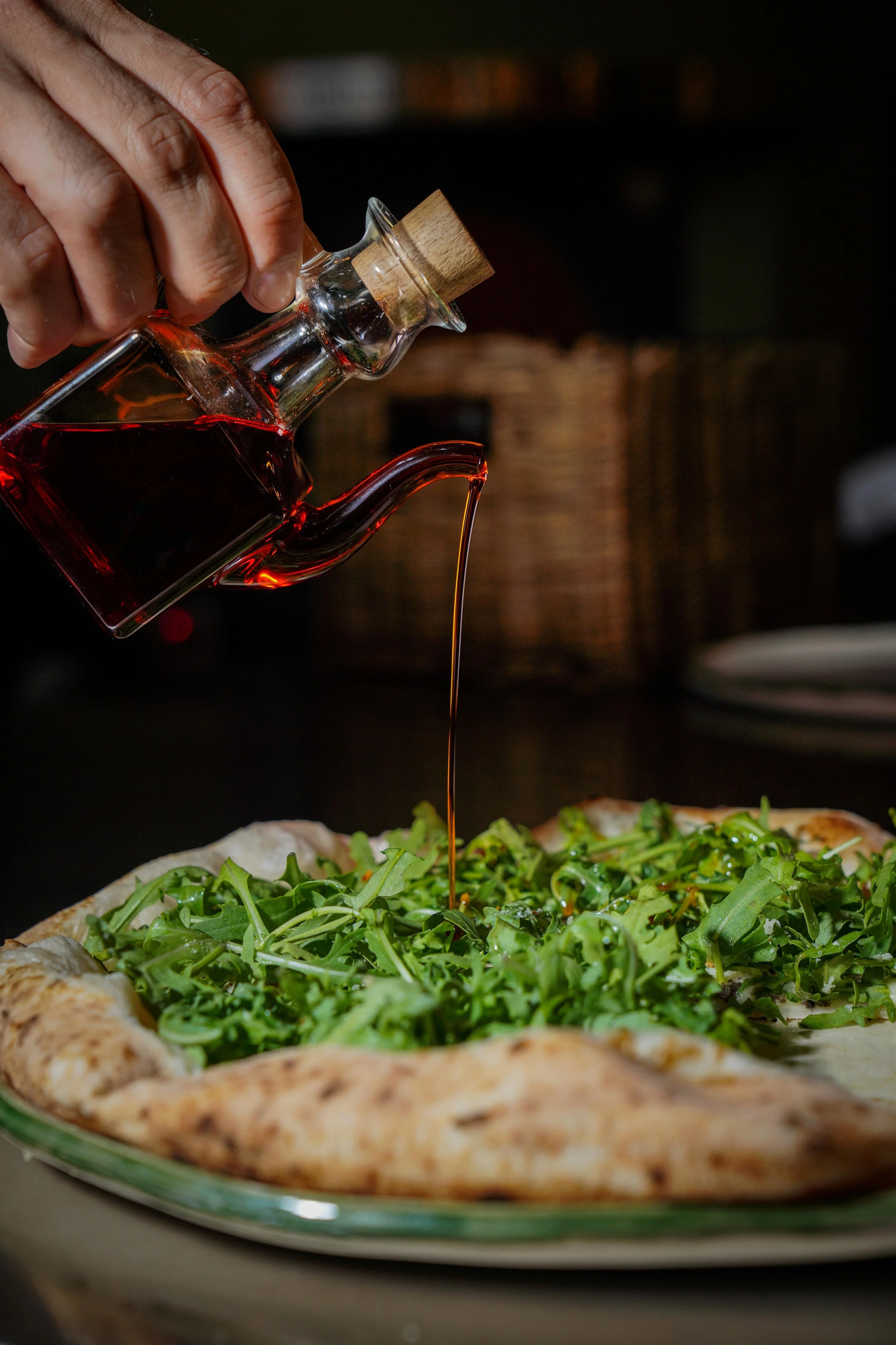 A hand pouring red wine over a pizza topped with arugula.
