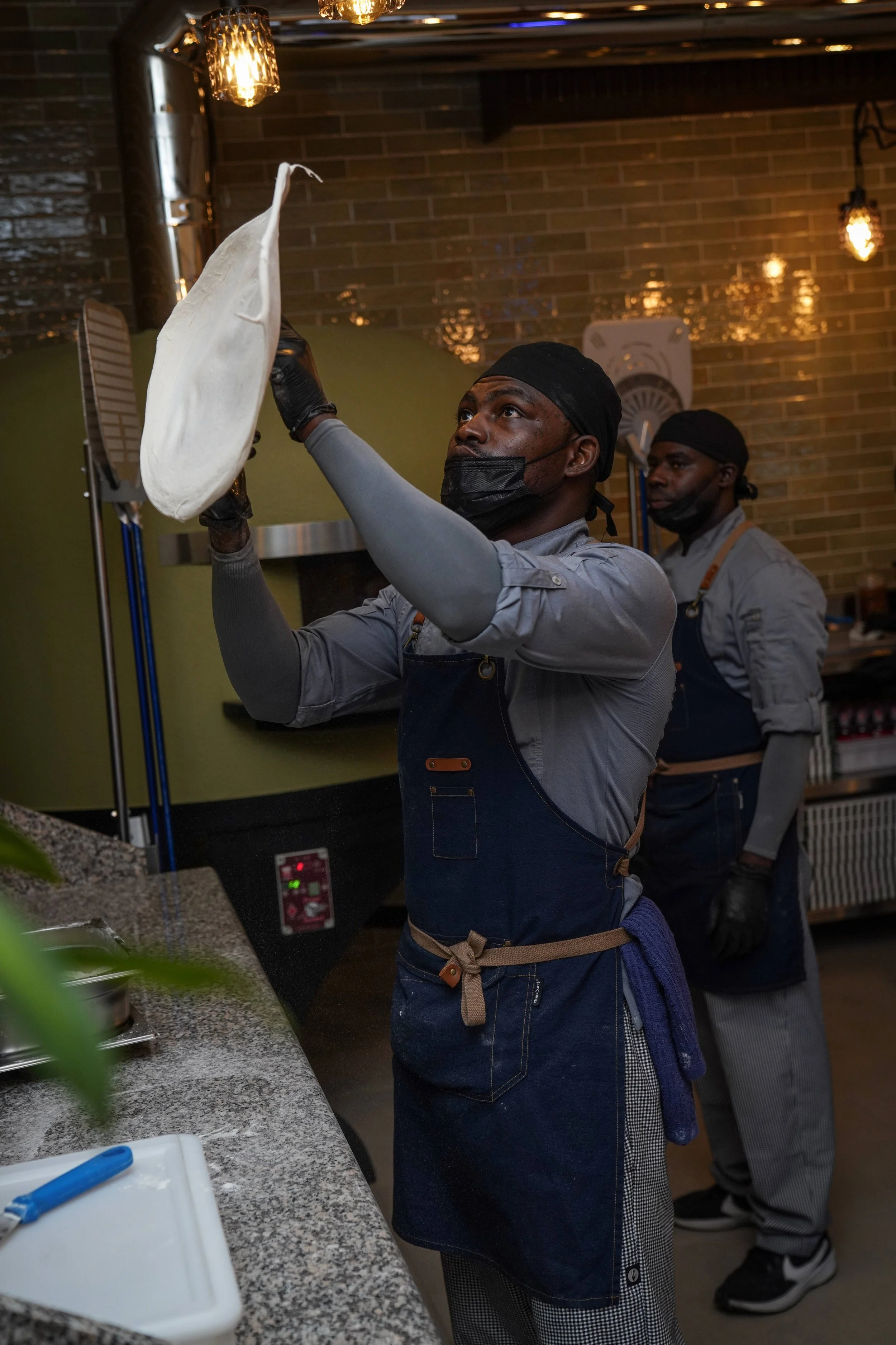 A man in a gray chef uniform and black apron is tossing a pizza dough in the air, while another man in similar attire stands behind him in a restaurant kitchen with warm lighting and brick walls.