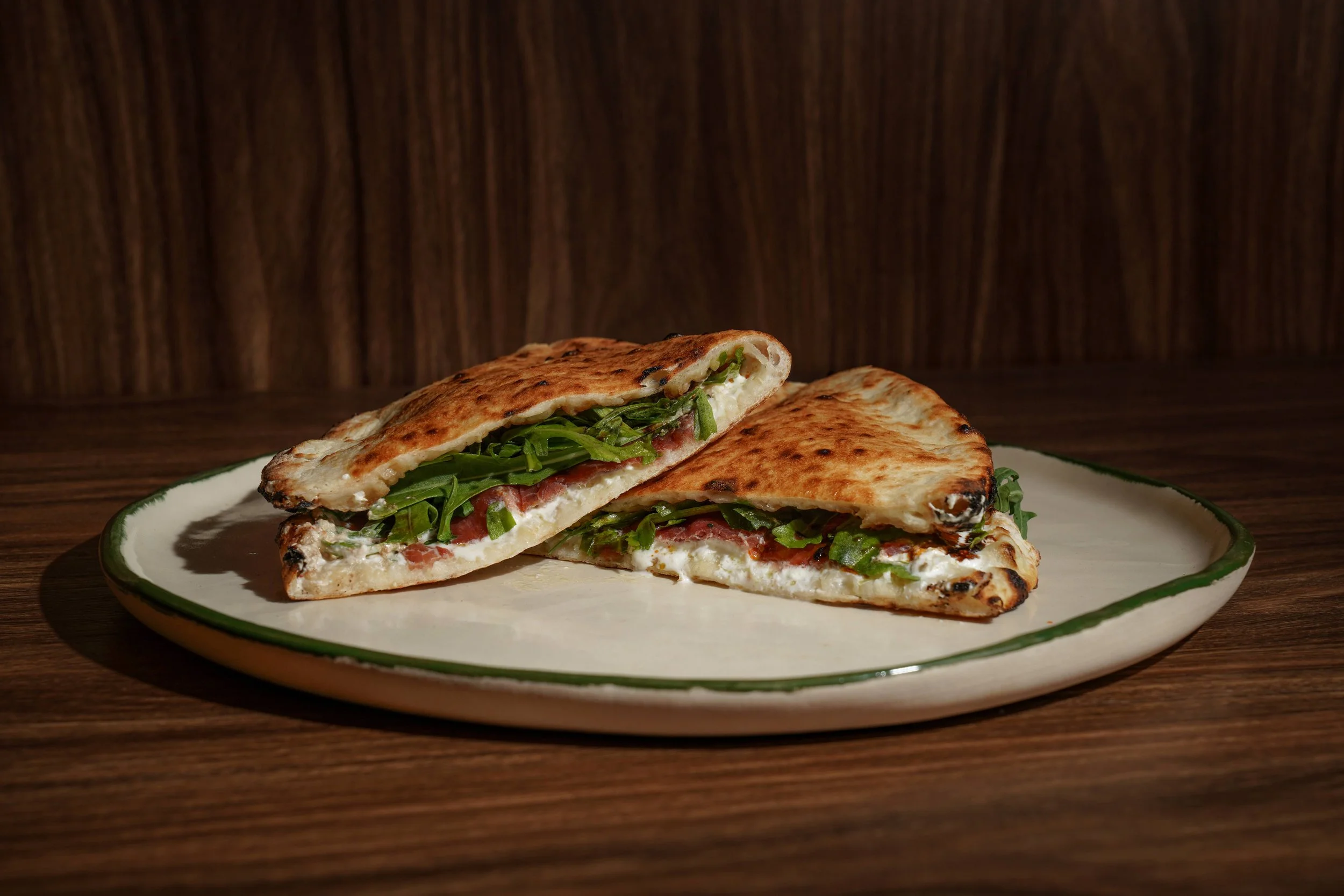 Half of a pizza with green vegetables and cheese on a white plate with a green rim, placed on a wooden surface.