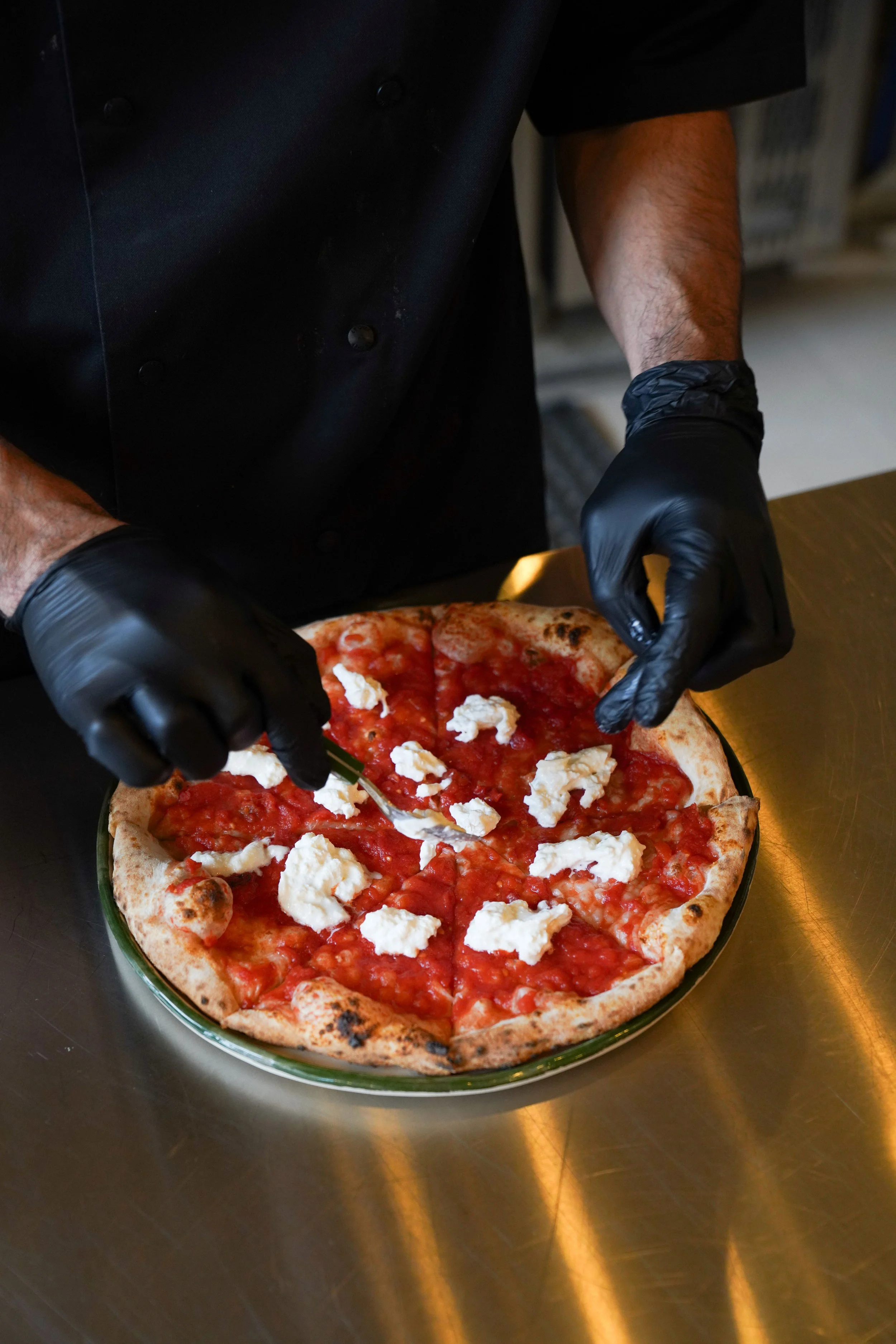 Chef wearing black gloves prepares to serve a pizza topped with tomato sauce and dollops of ricotta cheese on a green-bordered dish.