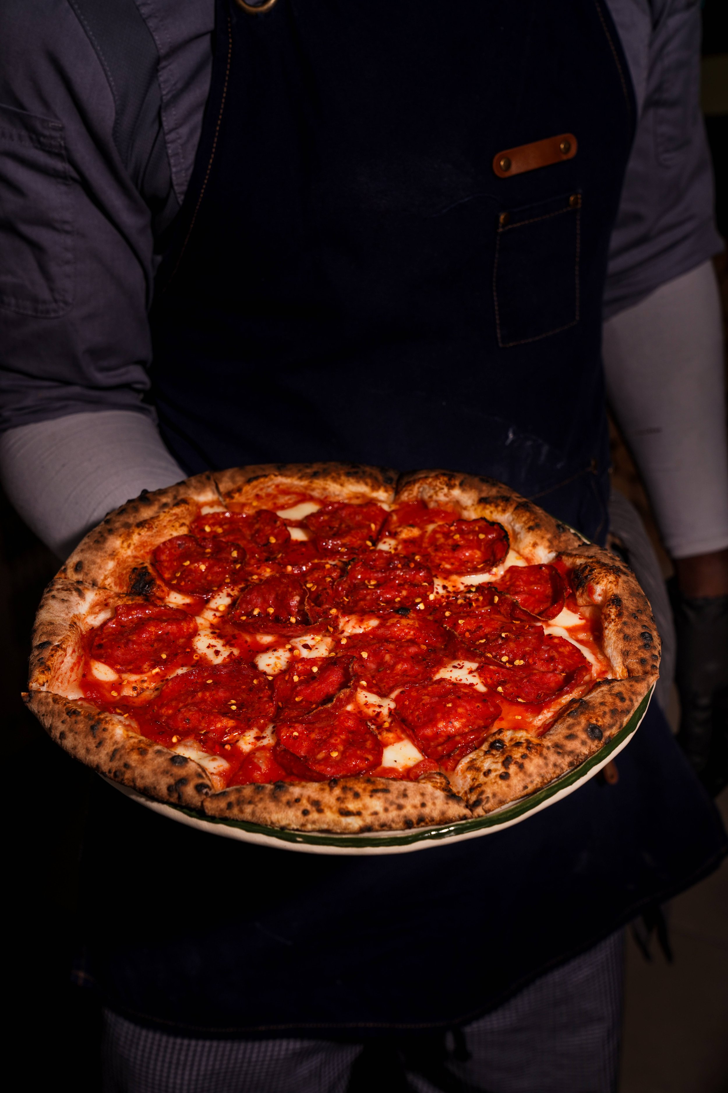 Person holding a pizza with pepperoni and cheese in a white bowl with green trim, wearing a dark apron.