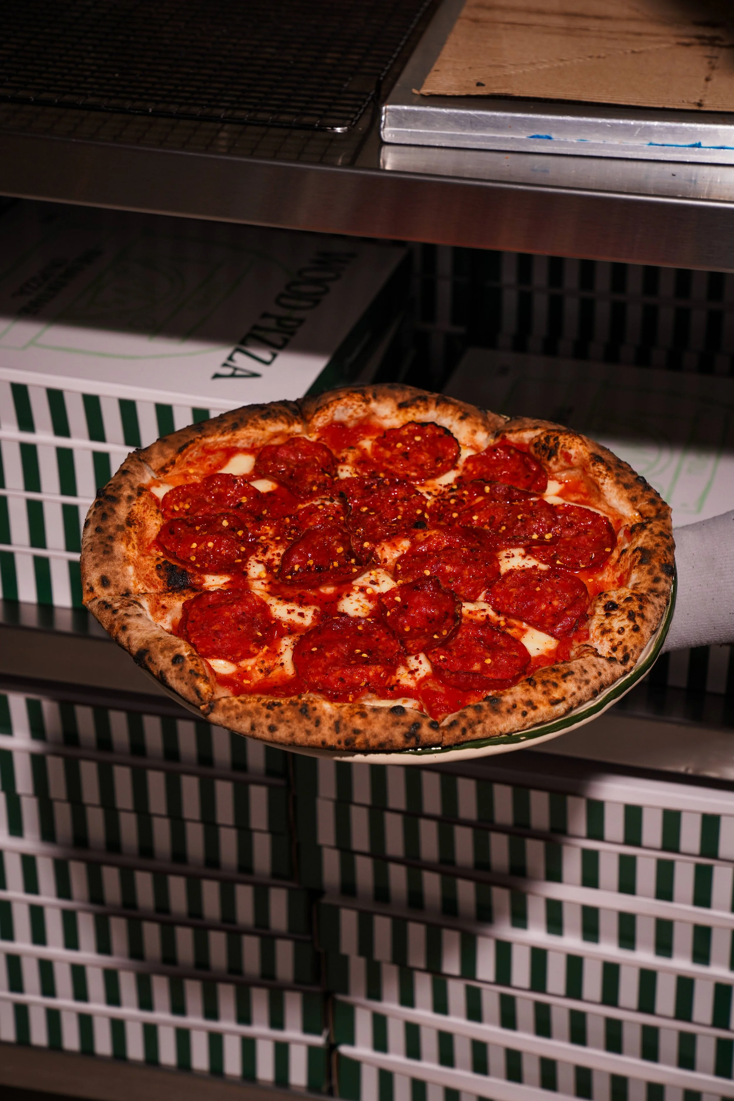 A pepperoni pizza in a green ceramic baking dish with a burnt crust, placed on a white wire shelf under a stainless steel oven or microwave.