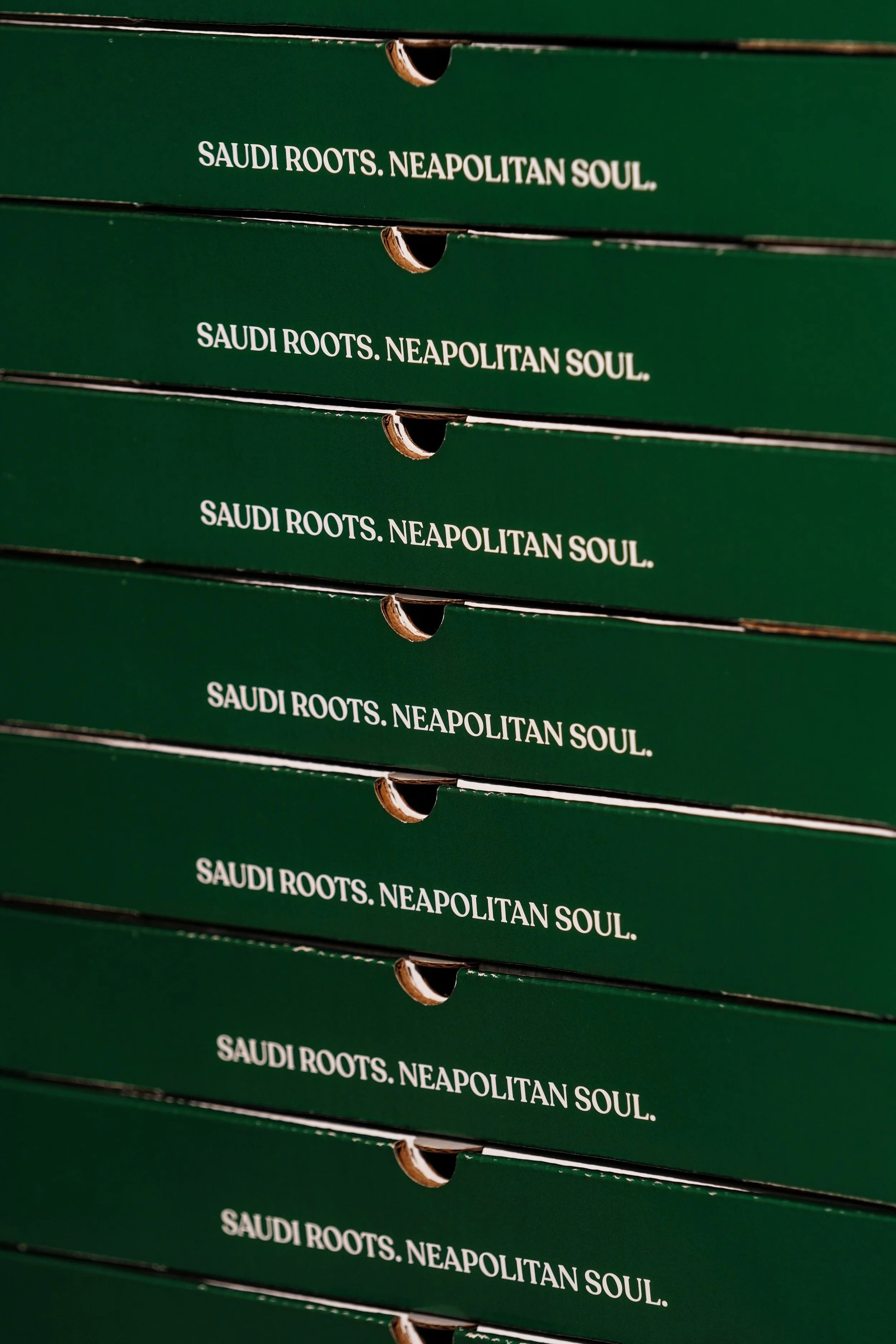 Close-up of green boxes with white text that reads 'SAUDI ROOTS. NEAPOLITAN SOUL.', stacked in a row.