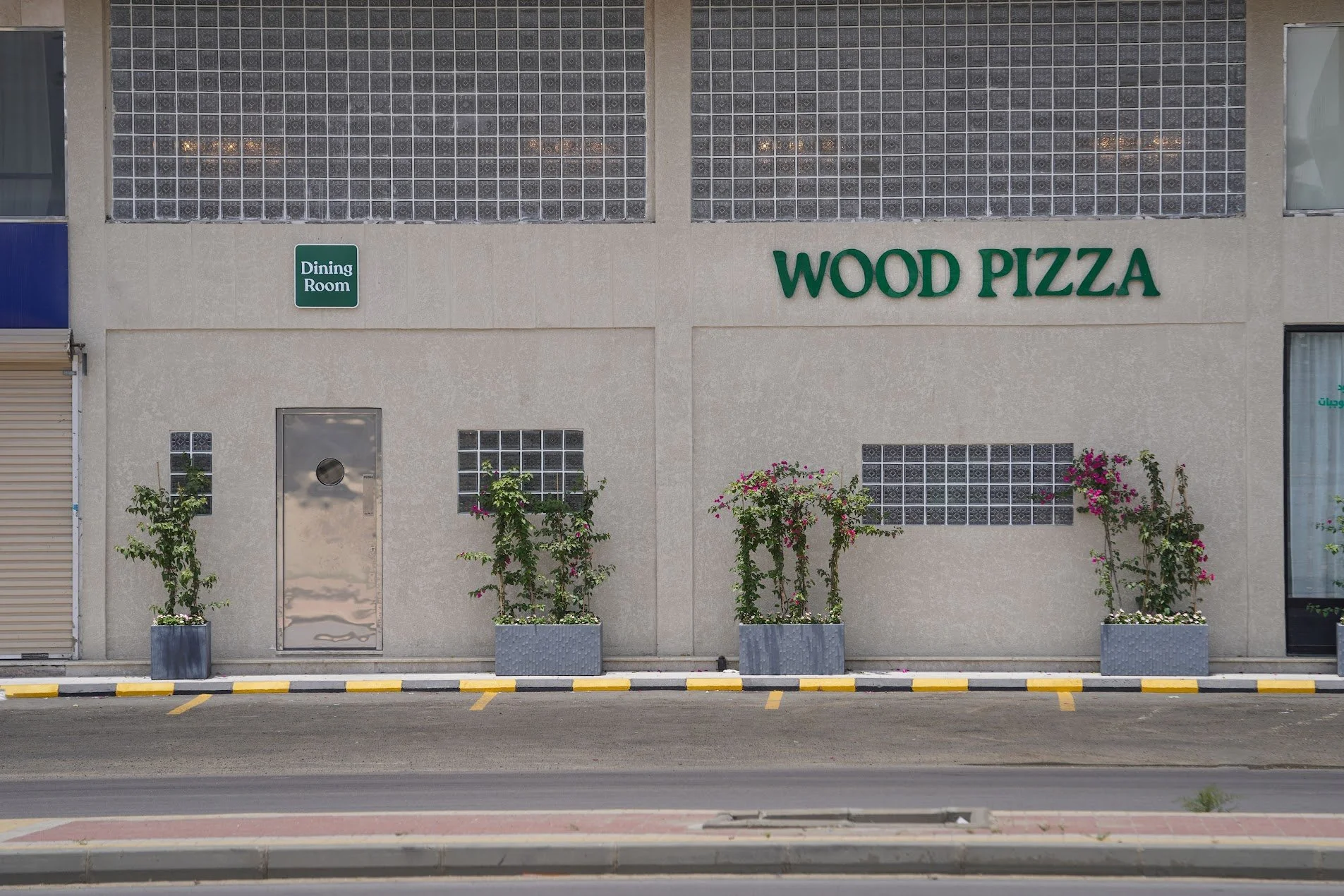 Facade of a building with a sign that reads 'WOOD PIZZA' and another sign indicating a dining room. There are four bushes with pink flowers in gray planters along the sidewalk, which also has yellow and black painted curbs and parking lanes.