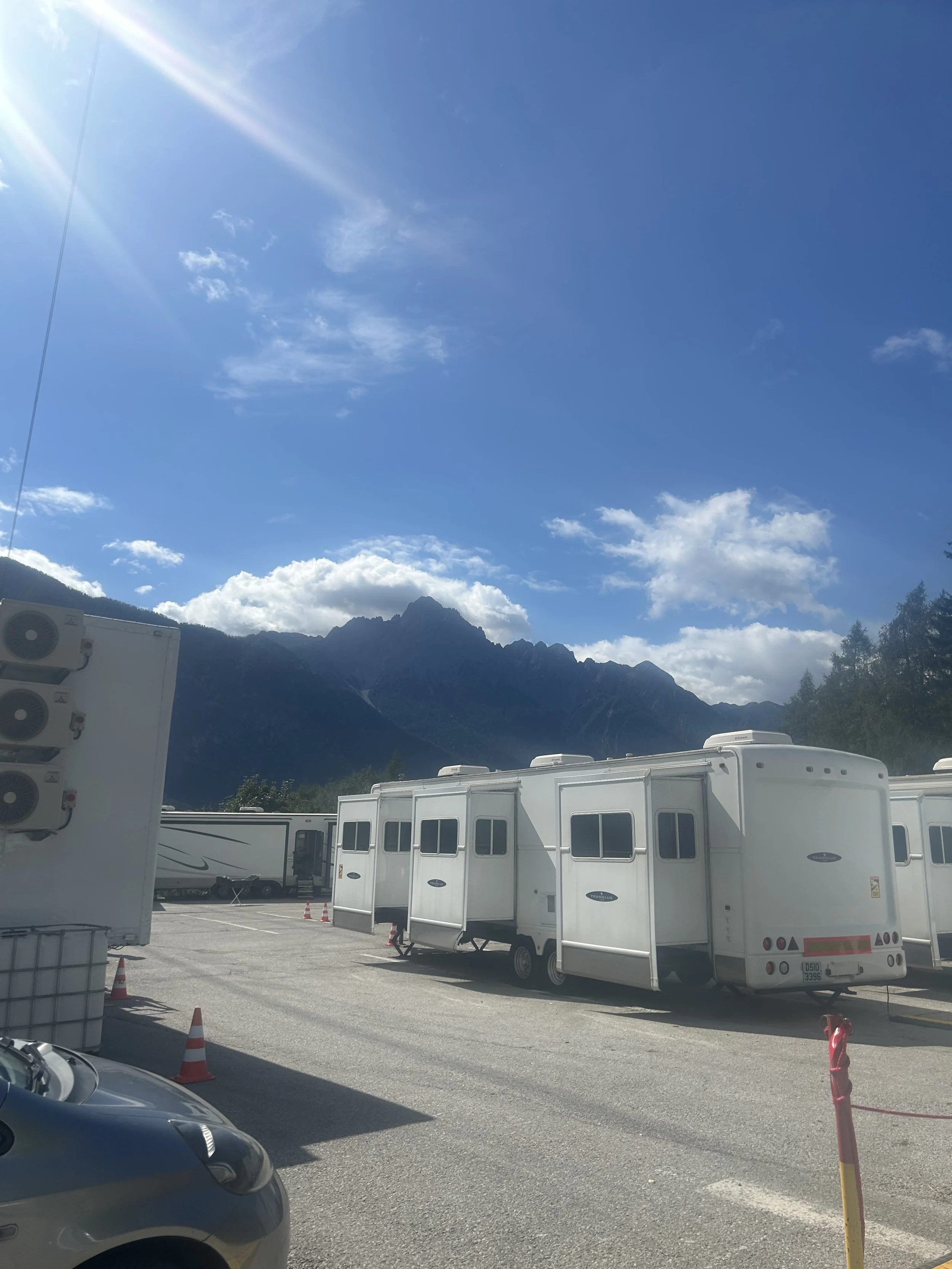 A parking lot with several white trailers and RVs, some with small windows, set against a backdrop of mountains, trees, and a blue sky with some clouds and sunlight.