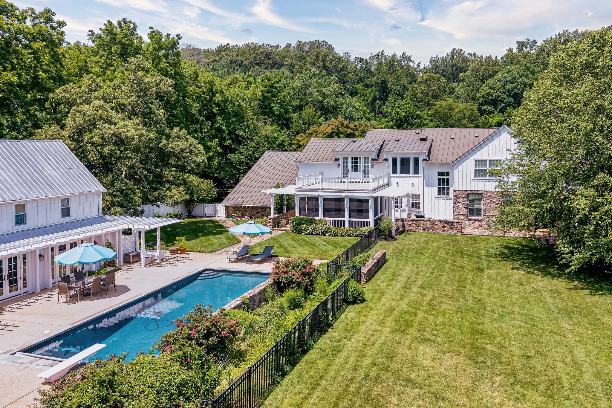 A backyard with a swimming pool, outdoor dining area with umbrellas, a lawn, and a white house with a brown roof surrounded by trees.