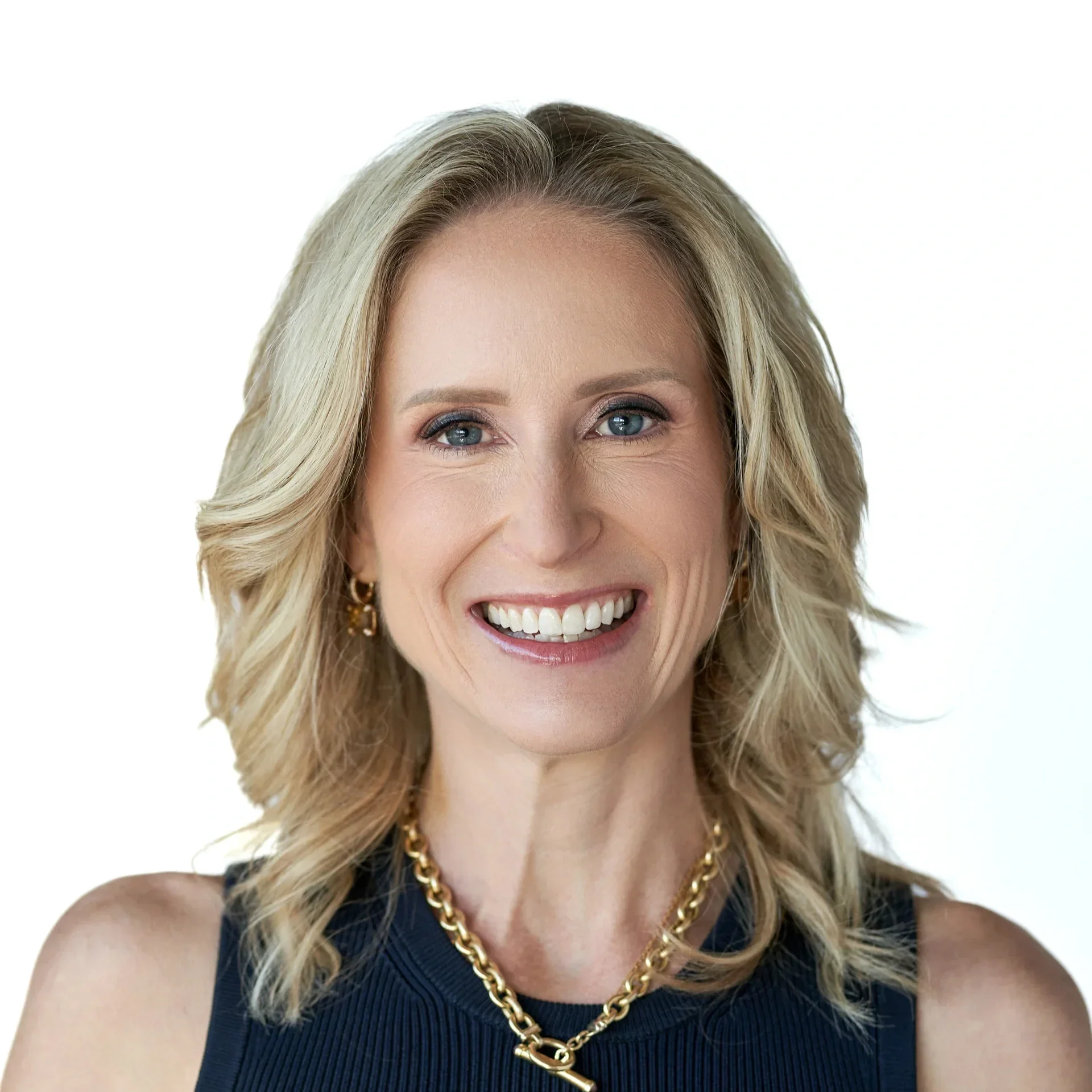 Close-up of smiling woman with blonde hair, blue eyes, wearing a black sleeveless top, gold chain necklace, and earrings, against a plain white background.