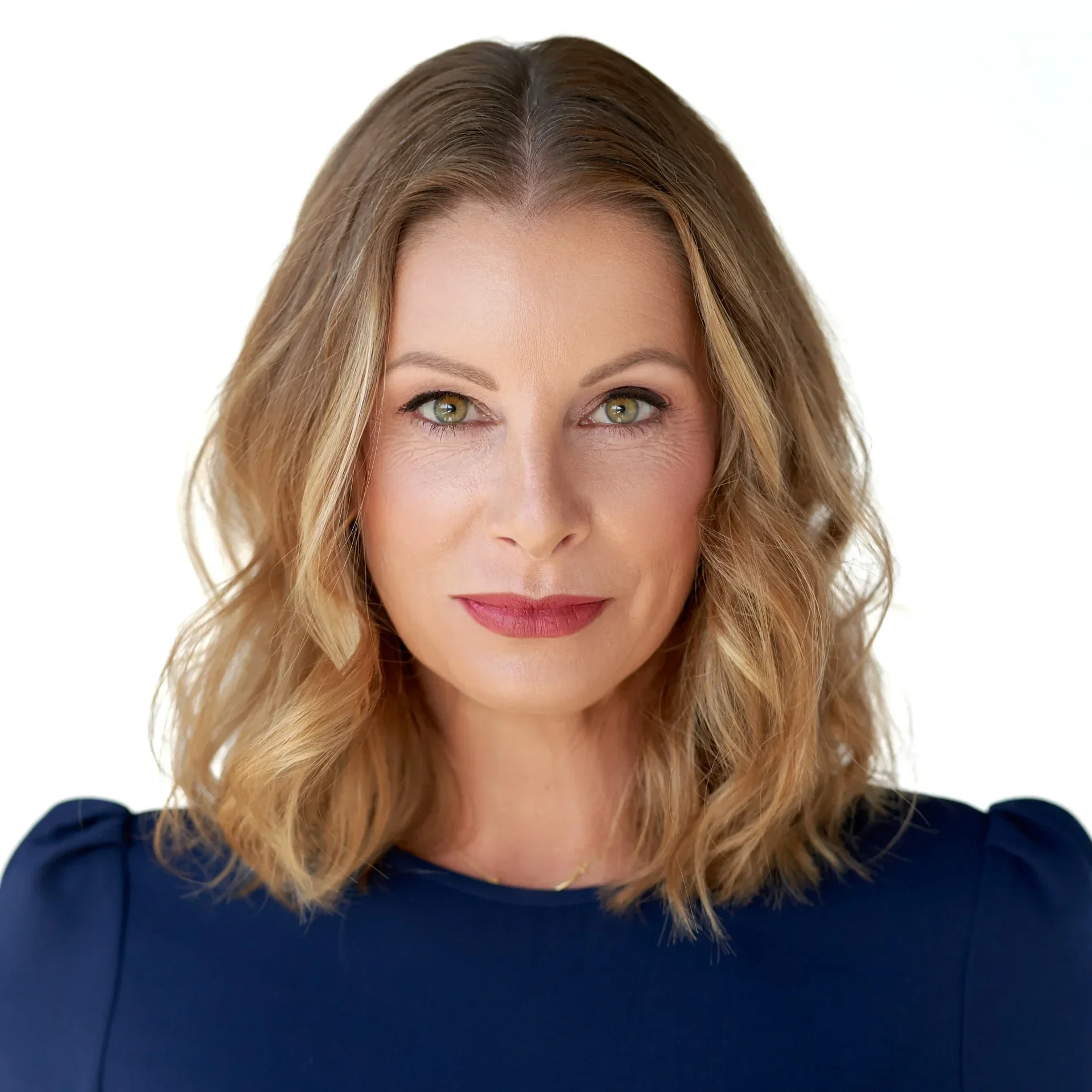 A woman with blonde, wavy hair and light skin, wearing a dark blue top, looking directly at the camera with a subtle smile, against a plain white background.