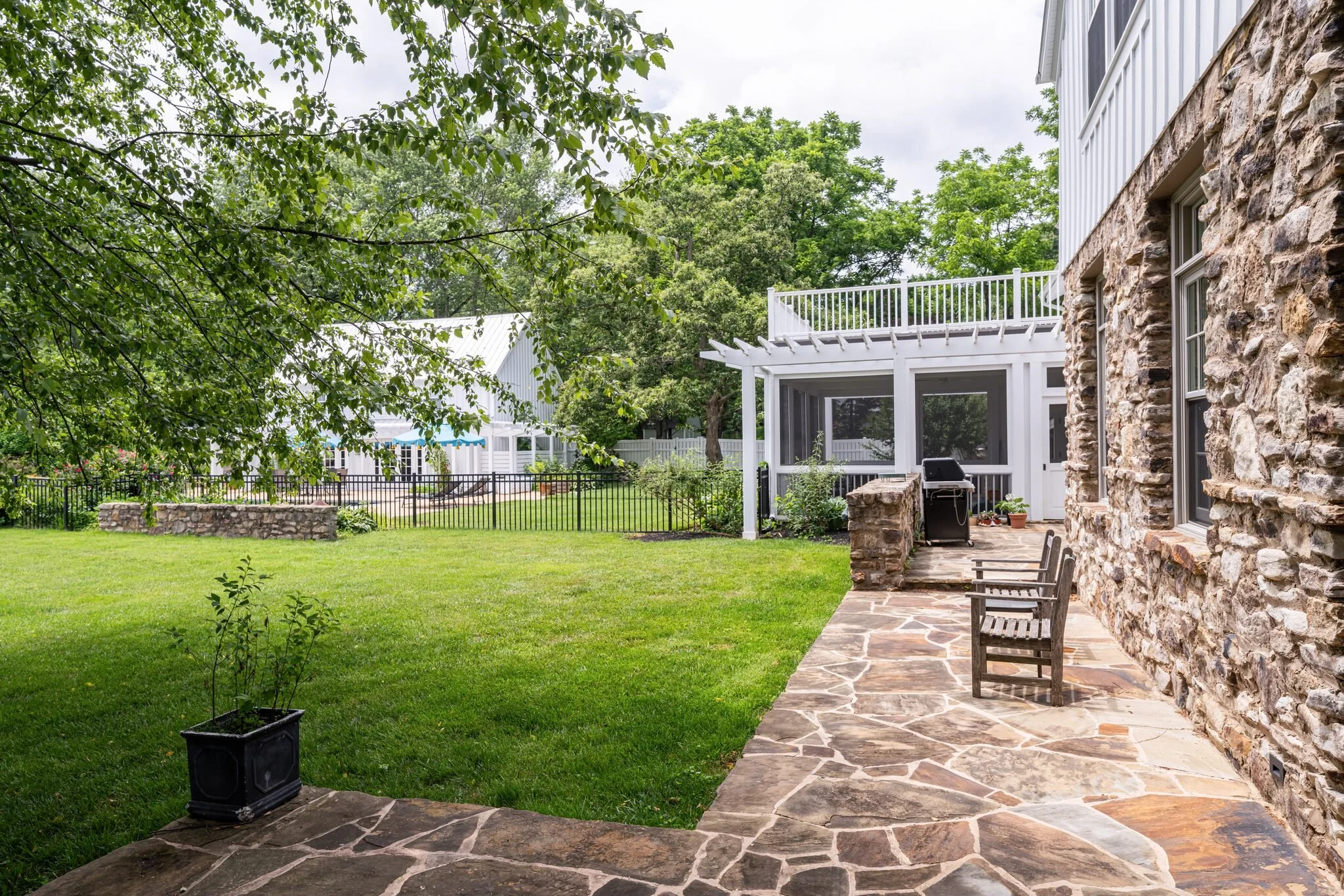 View of a backyard with a lush green lawn, stone patio, a stone house with large windows, a white wooden balcony, and surrounding trees