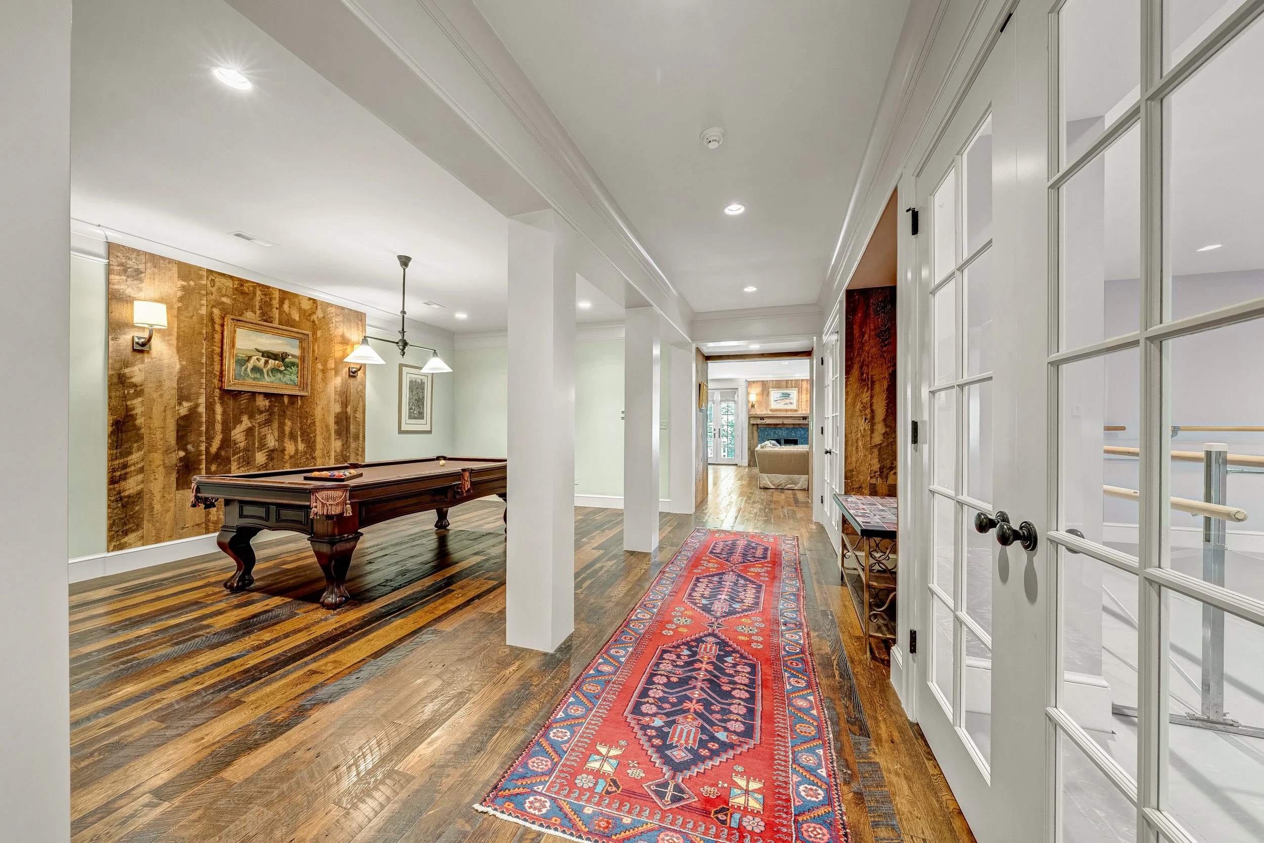 Interior view of a hallway and living area featuring a wooden pool table, a colorful runner rug, hardwood floors, and artwork on the walls, with natural light coming through windows and glass-paneled doors.