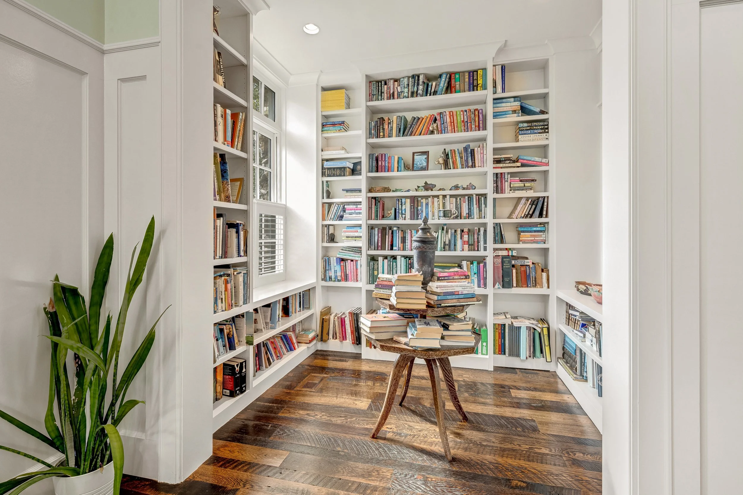 A cozy corner of a home library with white built-in bookshelves filled with various books. There is a wooden stool with a stack of books on it and a large decorative vase on top. A window with shutters lets in natural light. A tall green plant is in 