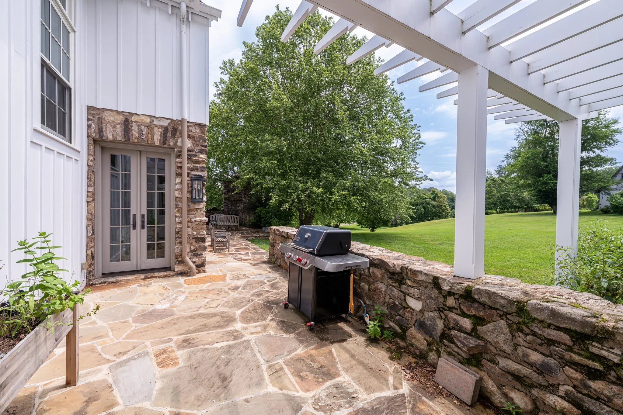 A backyard patio with a stone floor, a grill, a white house with large windows, a stone wall, and lush green trees in the background under a partly cloudy sky.