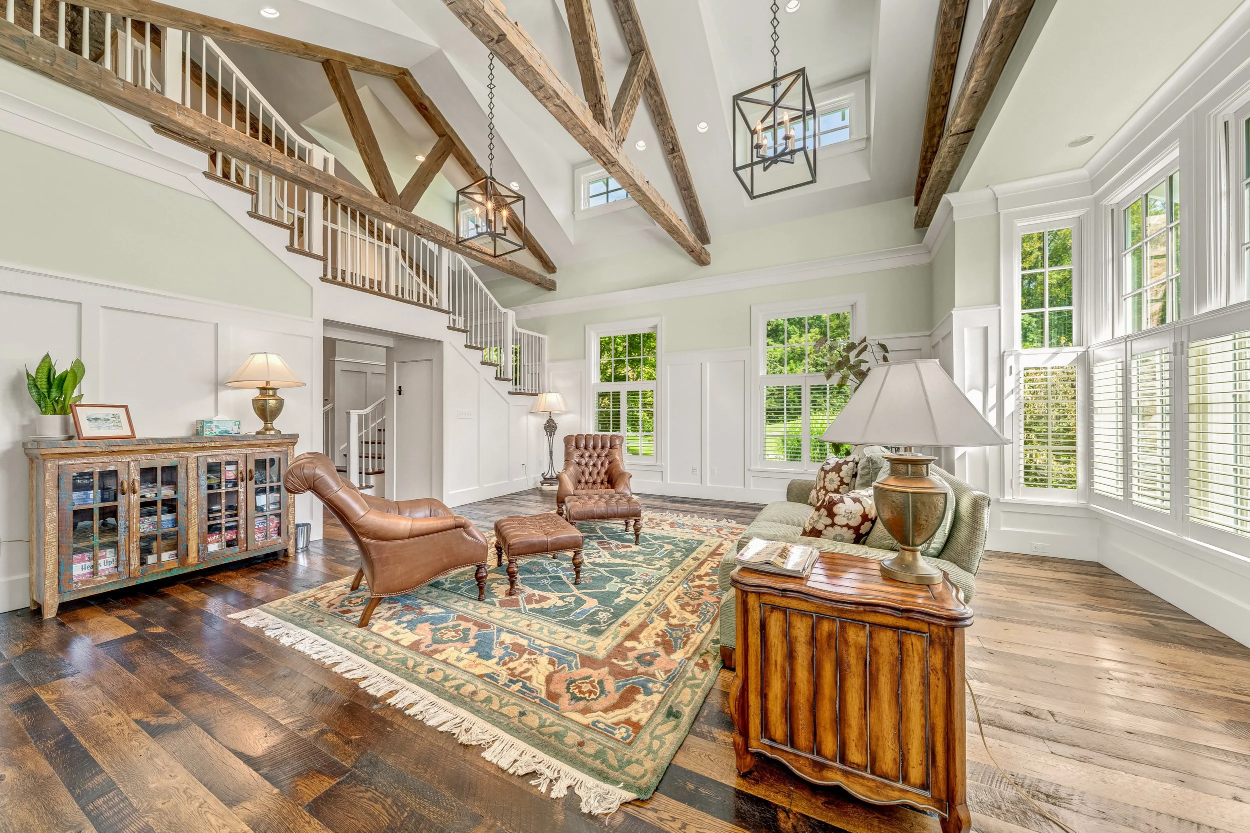 Living room with high vaulted ceiling featuring exposed wooden beams, large windows with white trim, and a mix of antique and modern furniture including leather armchairs, a green upholstered sofa, wooden side tables, and a patterned area rug.