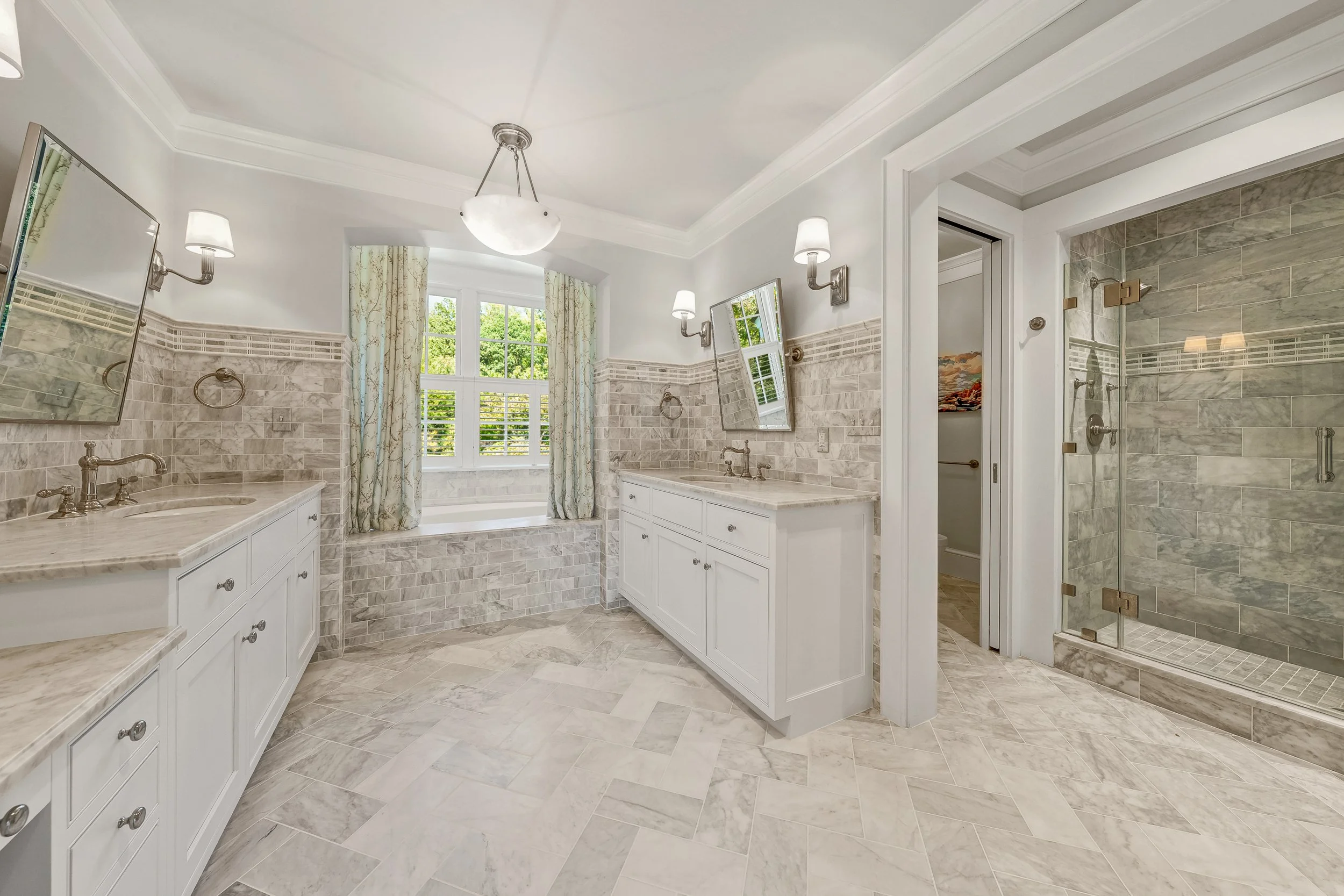 A spacious bathroom with white cabinetry, beige marble countertops, beige and gray marble tiles, a large window with plantation shutters, two wall-mounted lamps, and a glass-enclosed shower with matching marble tiles.