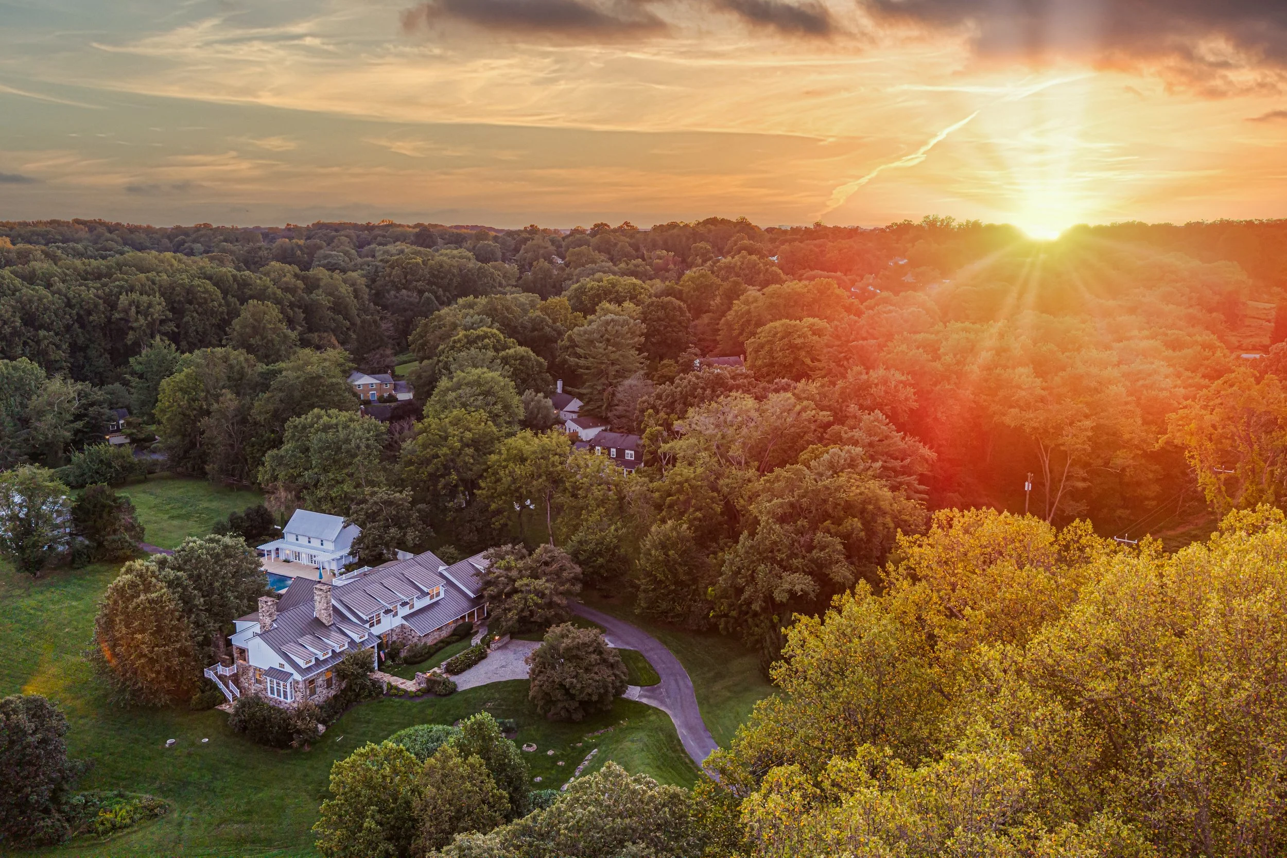 An aerial view of a suburban neighborhood during sunset, with a dense forest, large houses with landscaped yards, and a winding driveway.