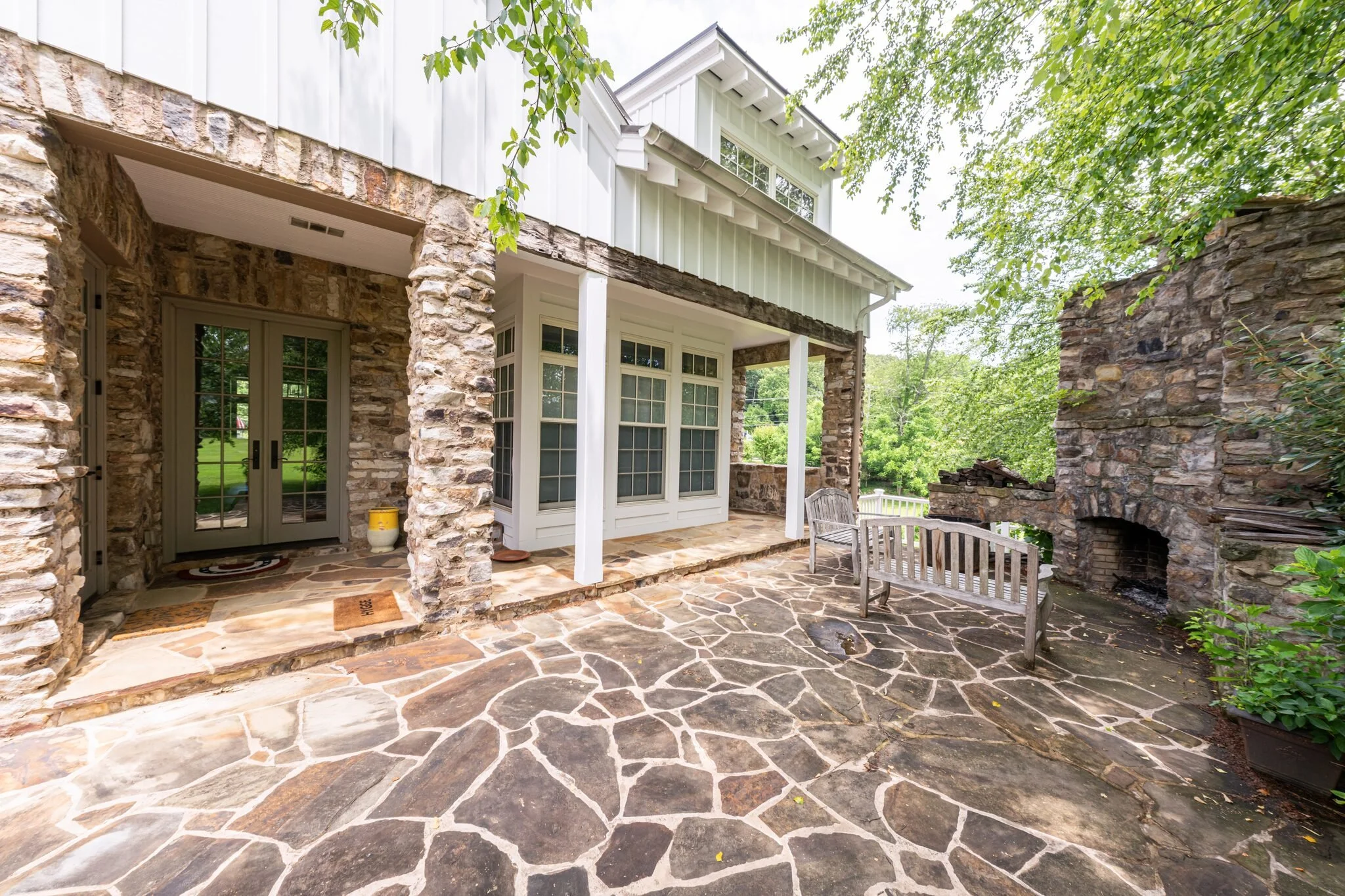 Back porch with stone flooring, stone columns, and a brick fireplace on the right, steps leading to a door with glass panes, surrounded by green trees and a white house exterior.