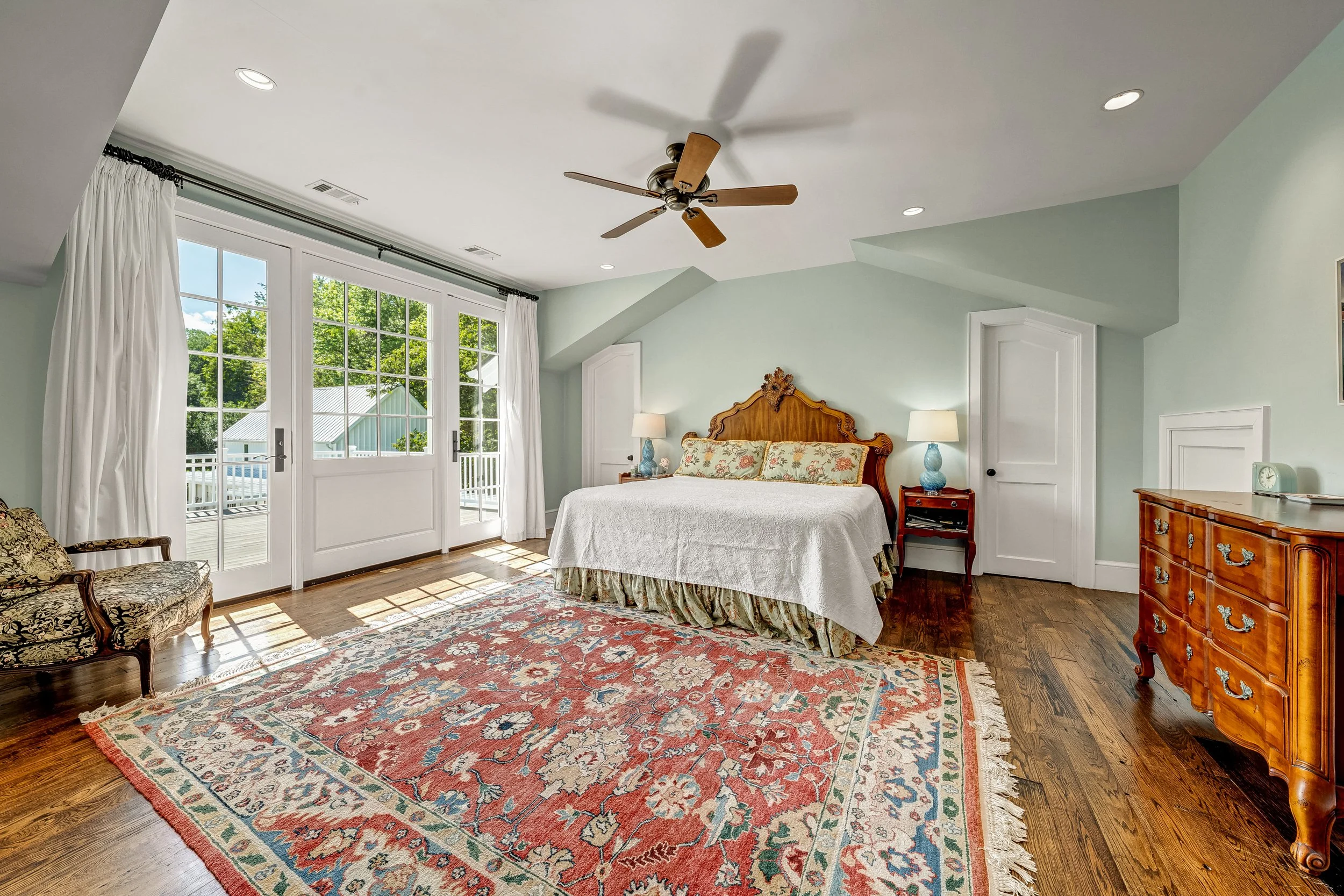A bedroom with a large bed, floral bedding, and a decorative wooden headboard. There are two nightstands with lamps, a sitting chair with ornate patterns, a colorful area rug, and large windows with white curtains letting in sunlight. The room has li