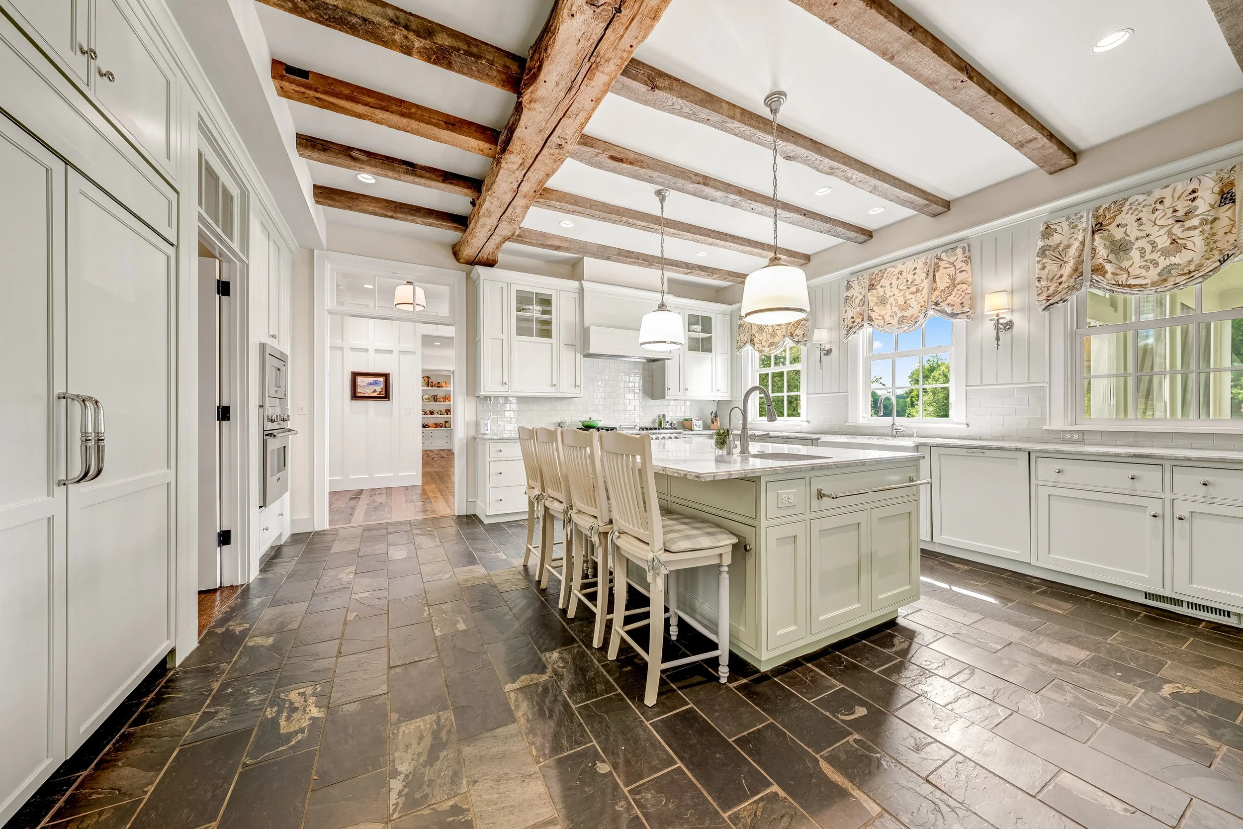A spacious kitchen with white cabinets, a large island, slate tile flooring, and exposed wooden ceiling beams. Large windows with floral valances let in natural light.
