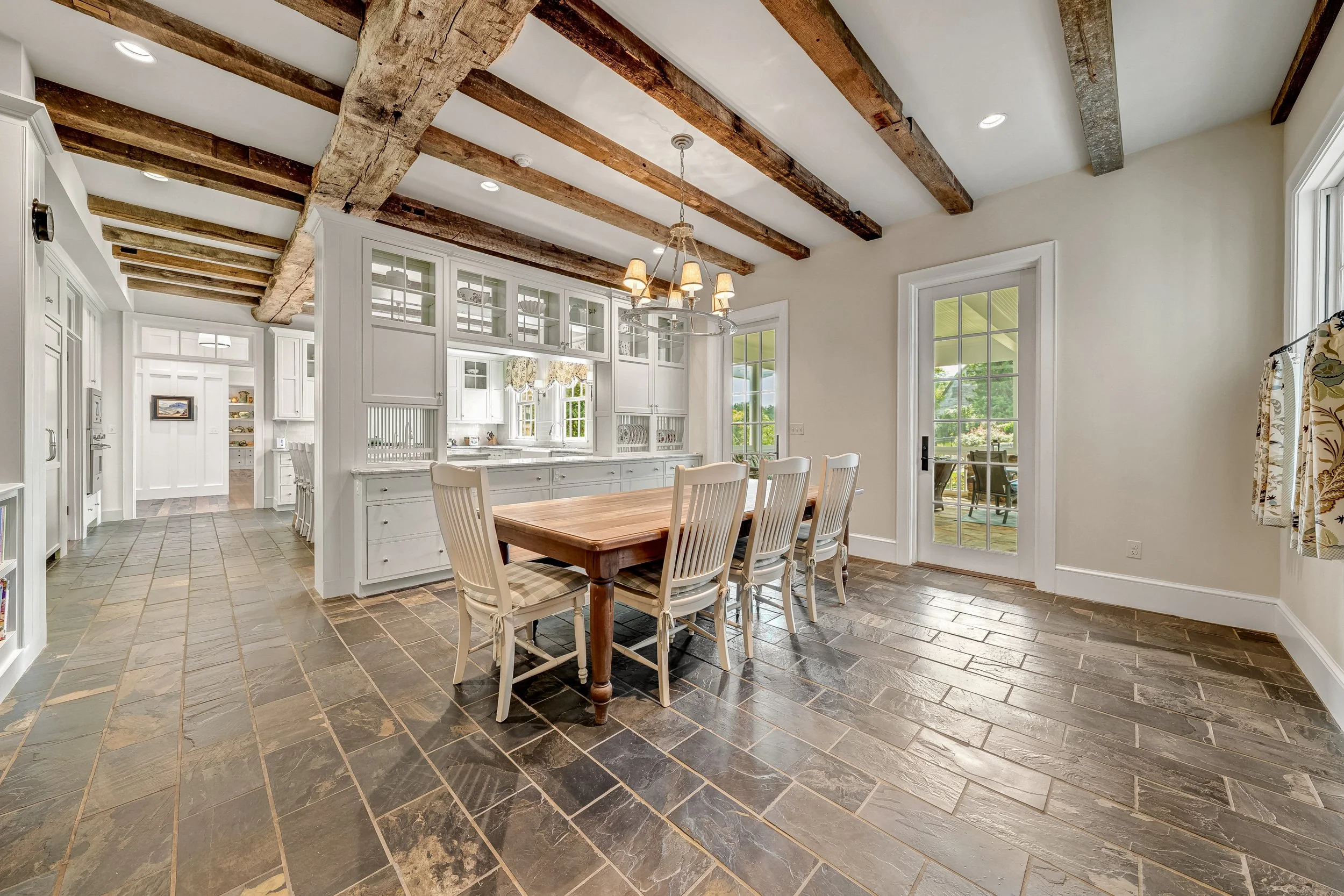 A spacious, bright kitchen and dining area with a rustic look. The ceiling has exposed wooden beams, and there are large windows and glass doors letting in natural light. The kitchen has white cabinets and a dining table with white chairs.