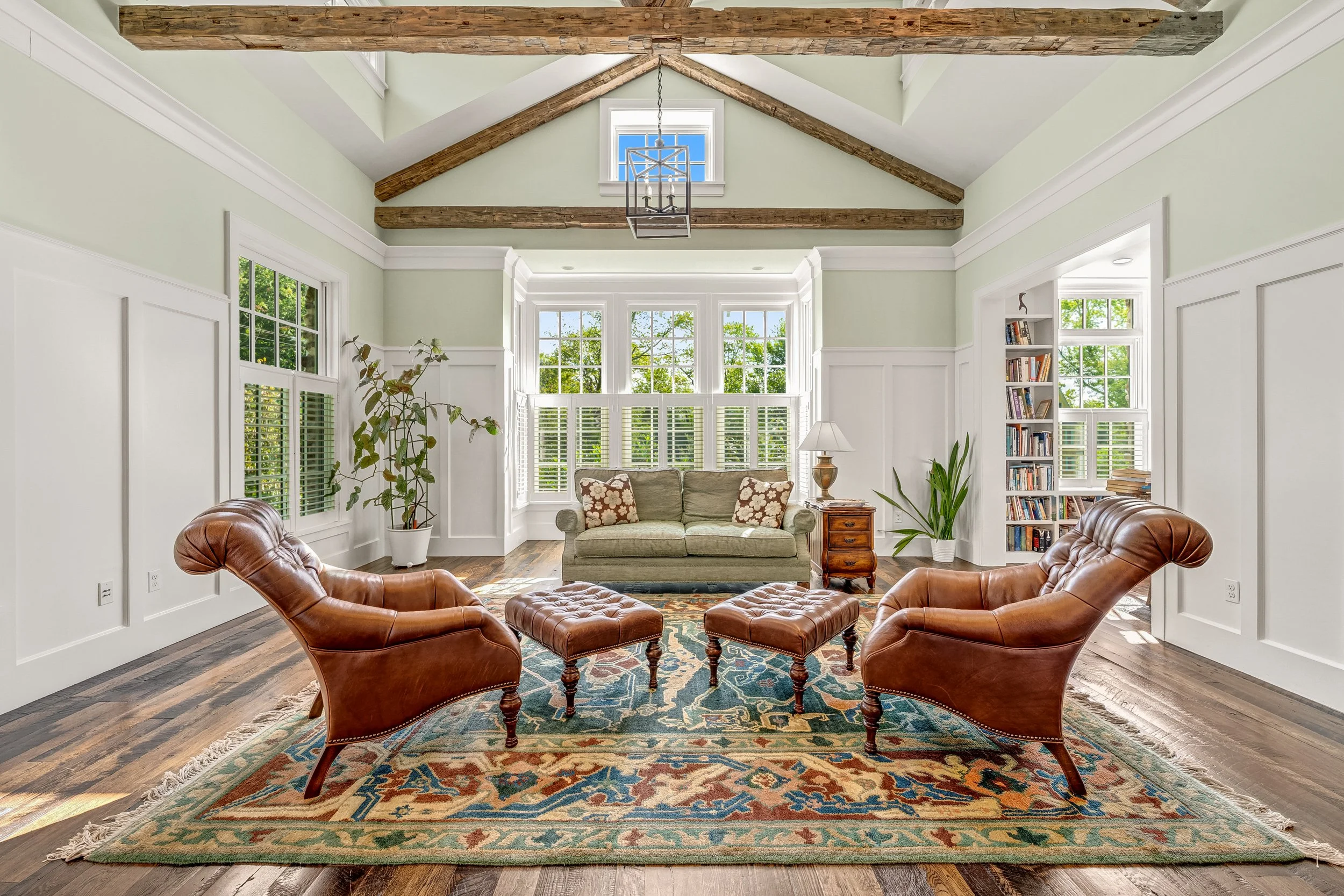 A bright sunlit living room with vaulted ceiling and exposed wooden beams, white paneled walls, hardwood floor, a green sofa with floral pillows, leather armchairs and matching footstools, a patterned rug, potted plants, large windows with white shut
