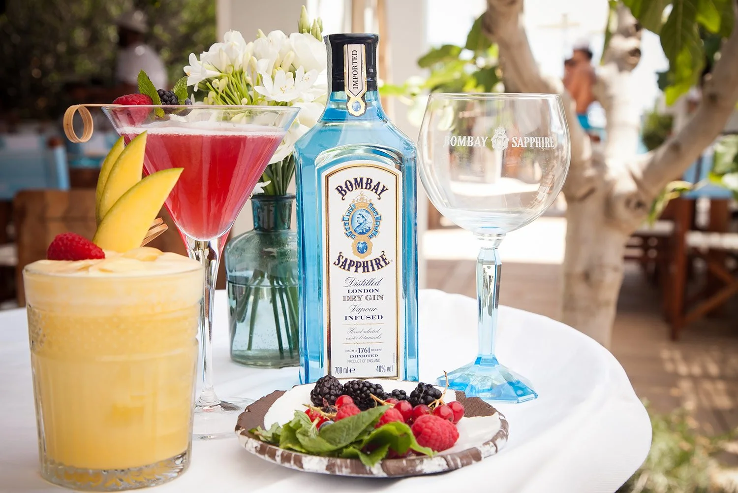 Bombay Sapphire Gin, colourful cocktails and a plated dish arranged on a sunlit outdoor restaurant table.