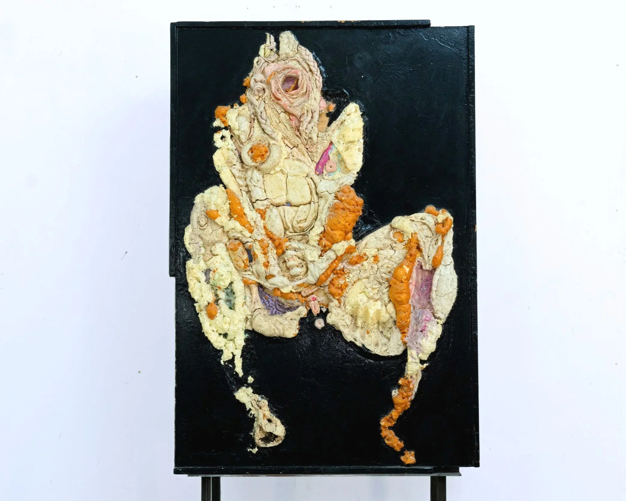An art piece depicting a human figure, made with a textured, layered material resembling clay or plaster, mounted on a black rectangular board. The figure seems to be seated with hands resting on the lap, with some orange and pink accents on the scul