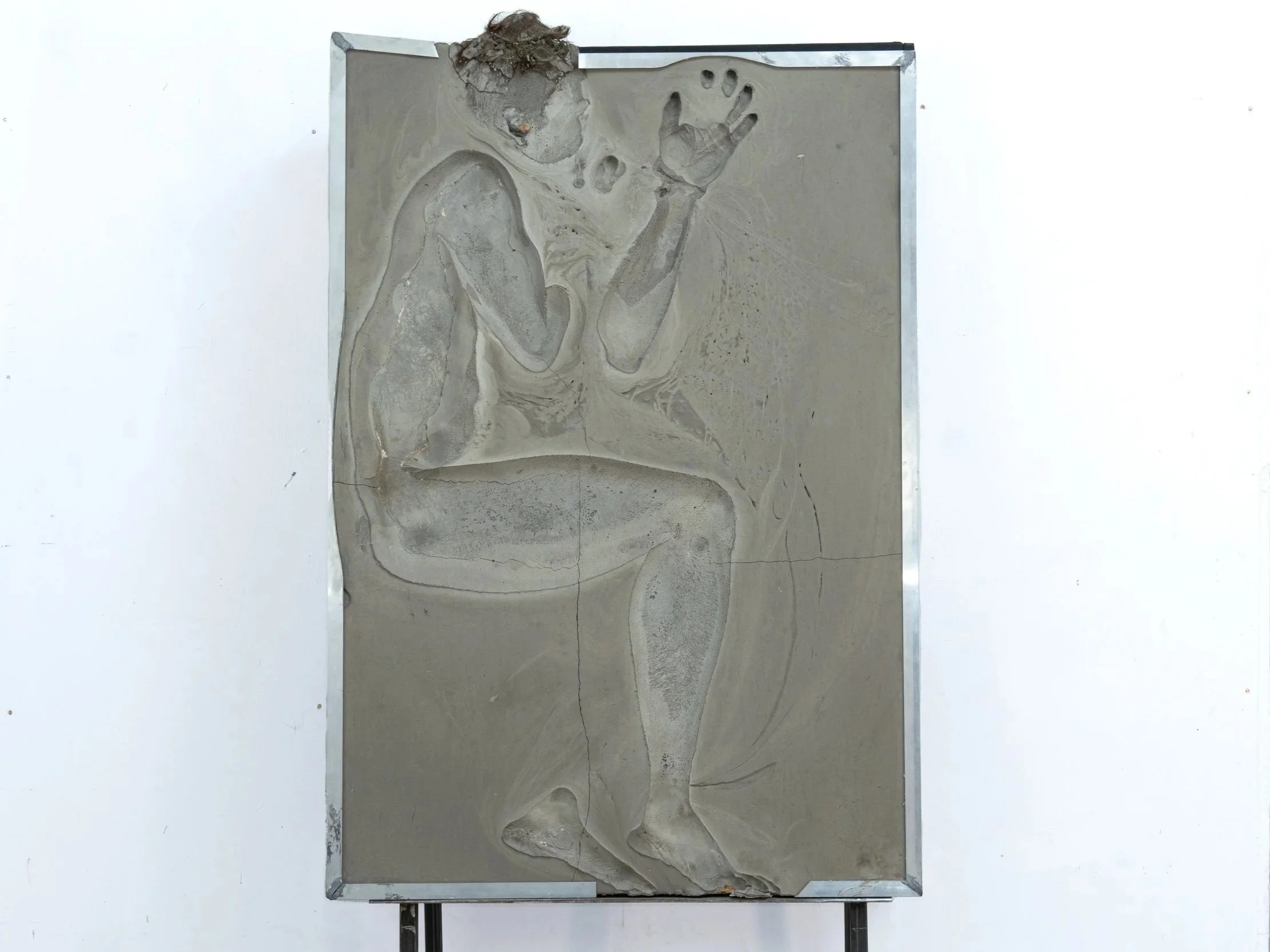 An artist's sculpture in a sandbox displays a detailed carving of a human figure with a raised hand, modeled in gray material surrounded by a metallic frame, on a white background.