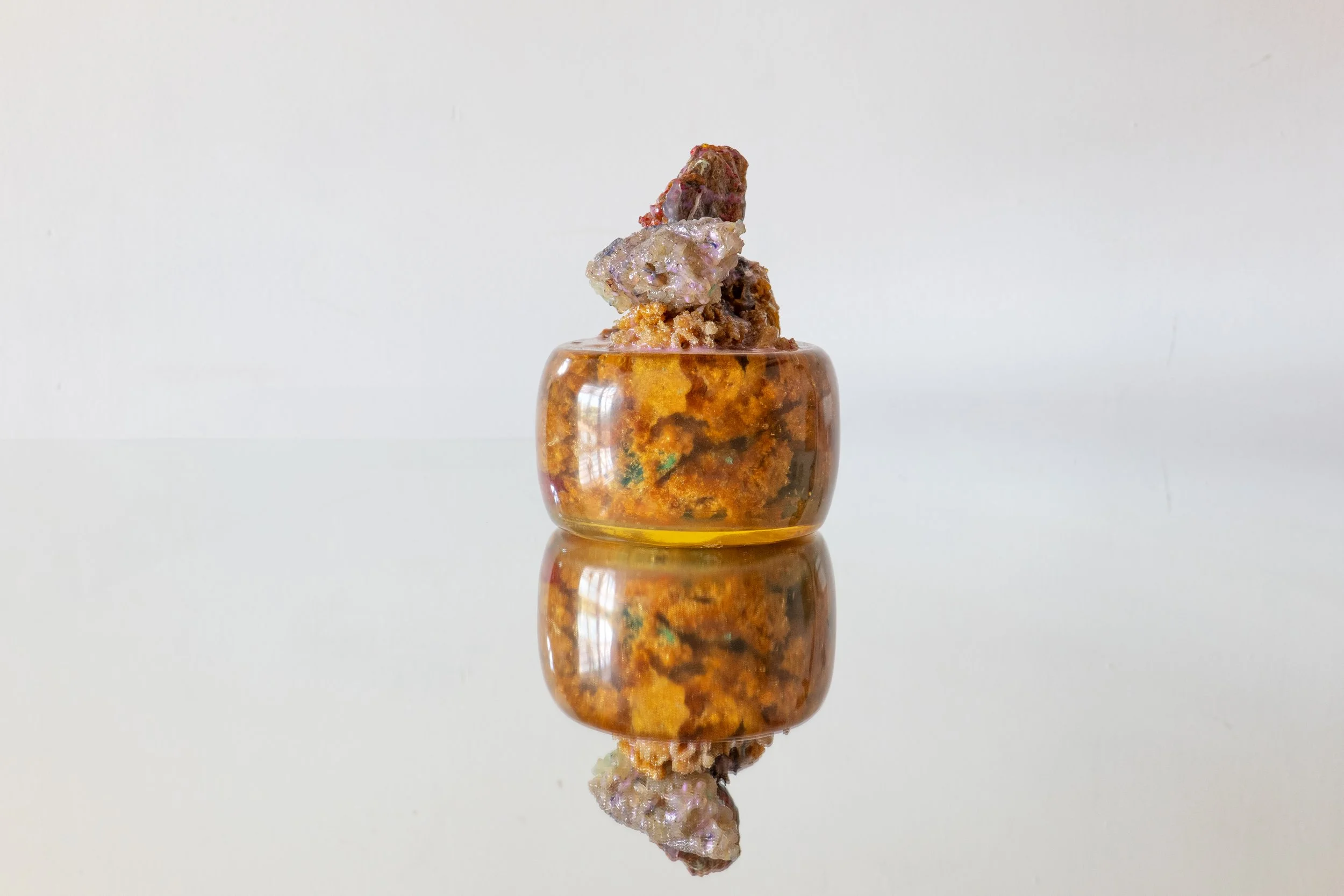 A small, round, amber-colored glass container with a textured, multicolored sculpture on top, set against a plain white background with its reflection visible on the surface beneath.