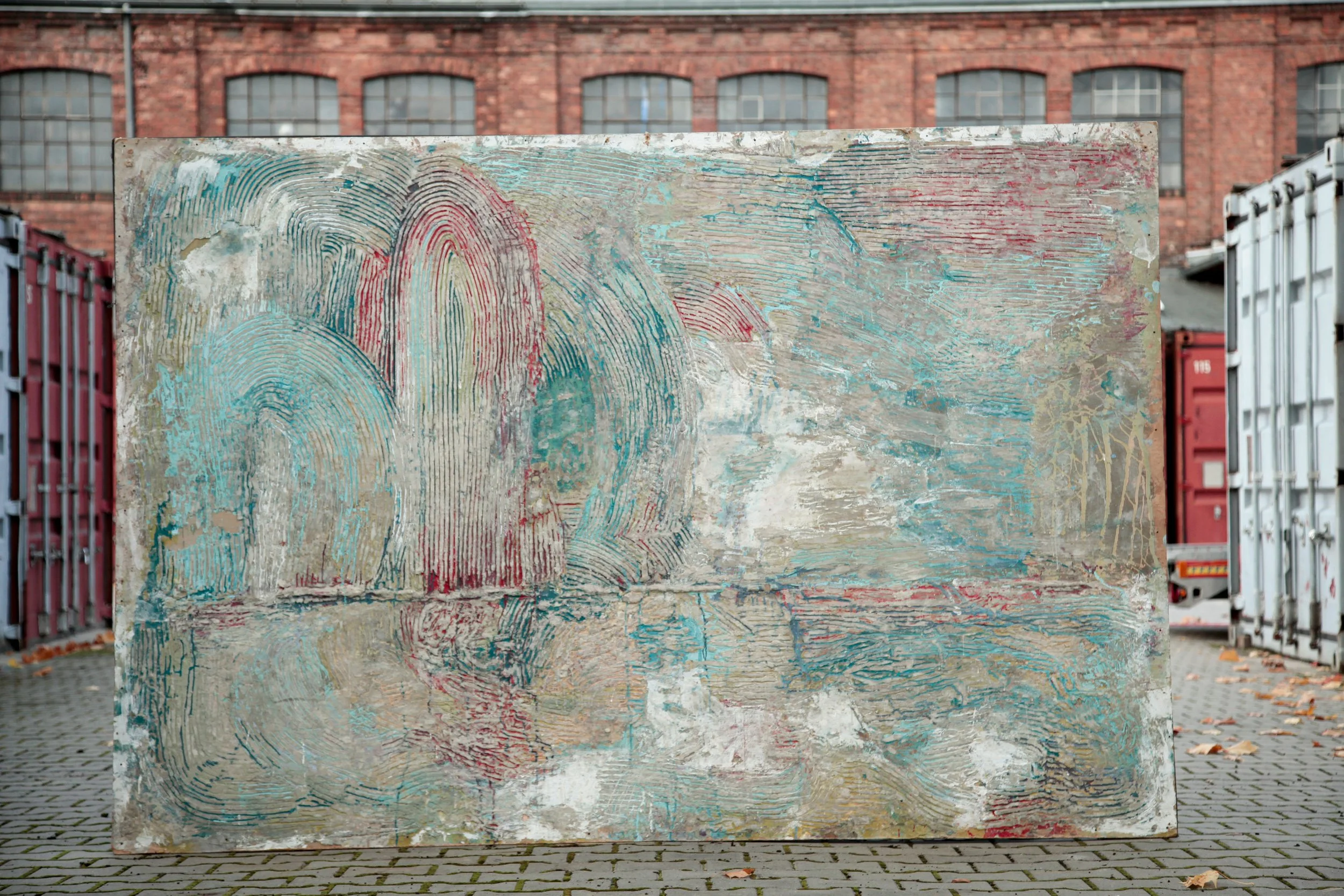A large weathered metal panel with colorful abstract, textured patterns featuring curved lines in red, blue, and beige, outdoors on a cobblestone surface with a brick building and shipping containers in the background.