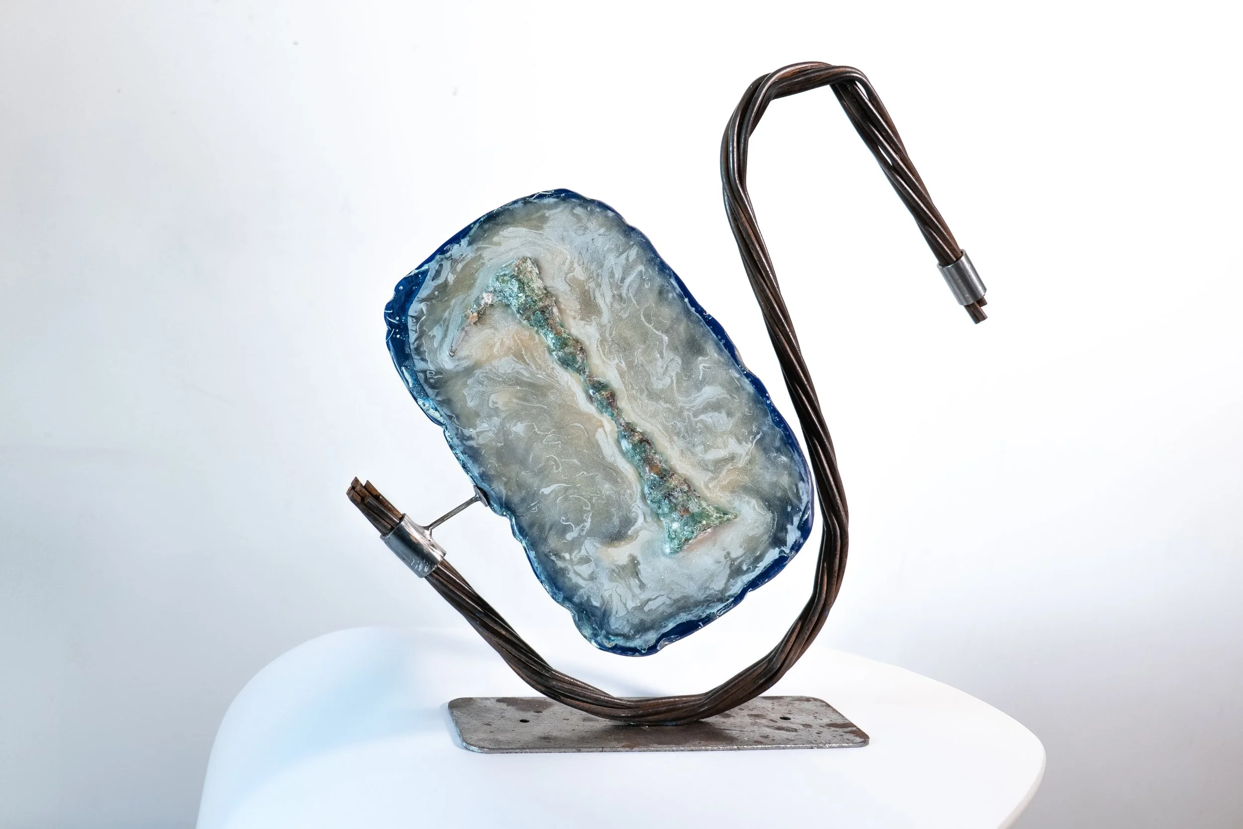 A piece of polished agate with blue, cream, and green colors mounted on a metal stand. It is connected to a brown wire with metal connectors on each end for electrical use.