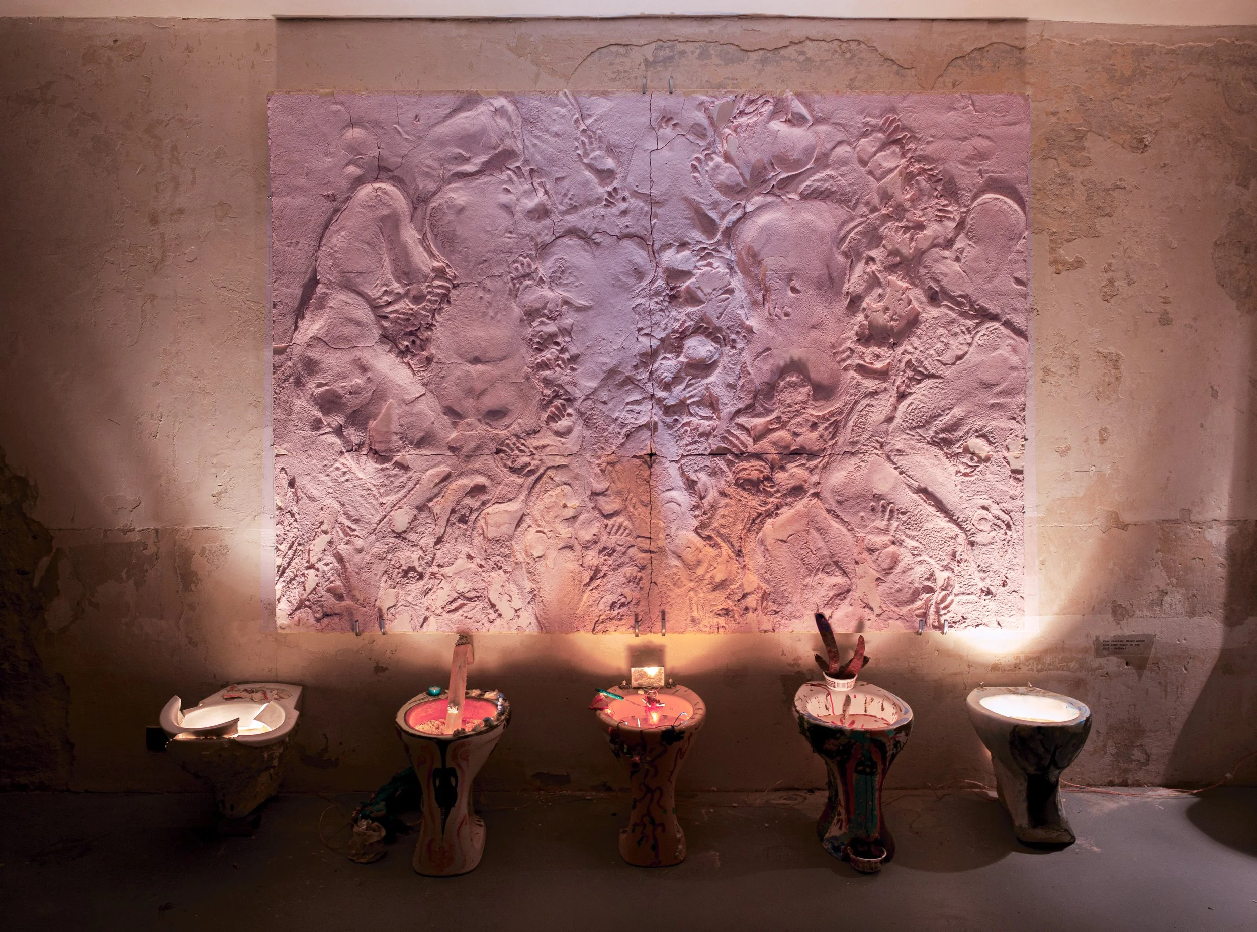 An art exhibit featuring a textured pink wall with fossil-like imprints and art installation bowls below it, illuminated with focused lighting.