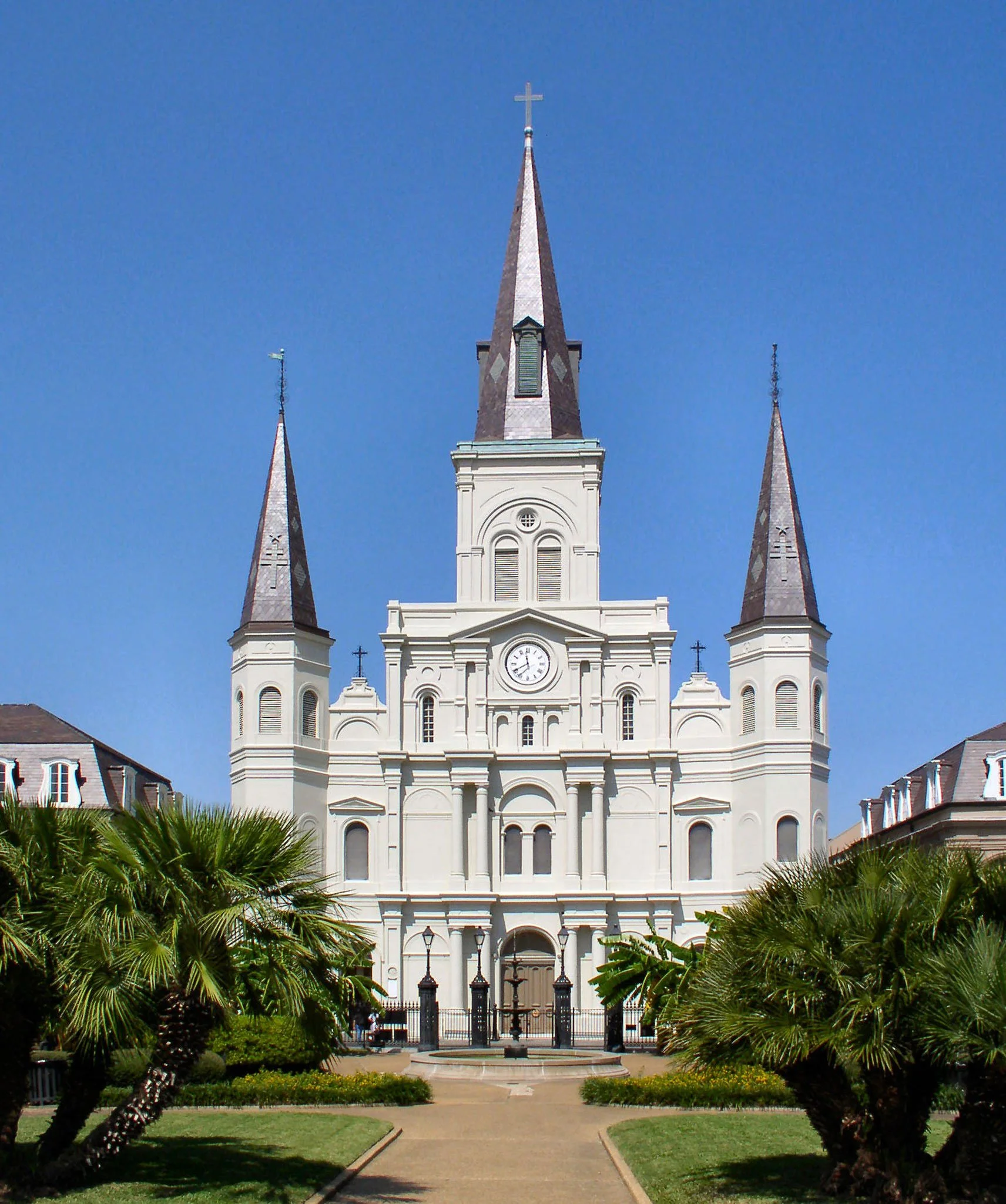 Front view of a white church with a clock on its facade, three steeples with crosses on top, surrounded by palm trees and greenery under a clear blue sky.