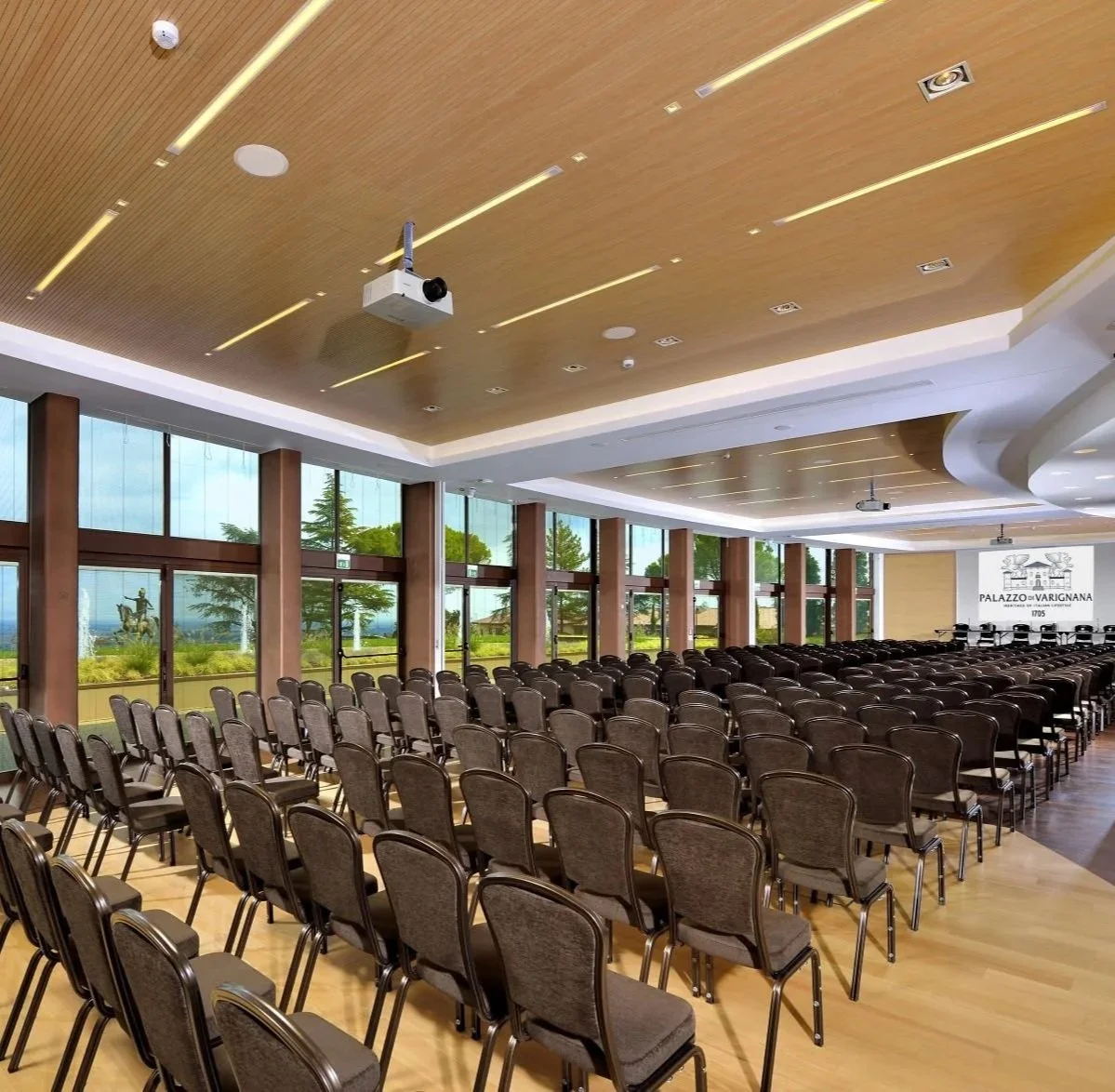 Varignana Summer Institute by Medevise Consulting. Conference room with rows of chairs, large windows showing trees outside, and a screen displaying 'Palazzo di Varignana 1705'.