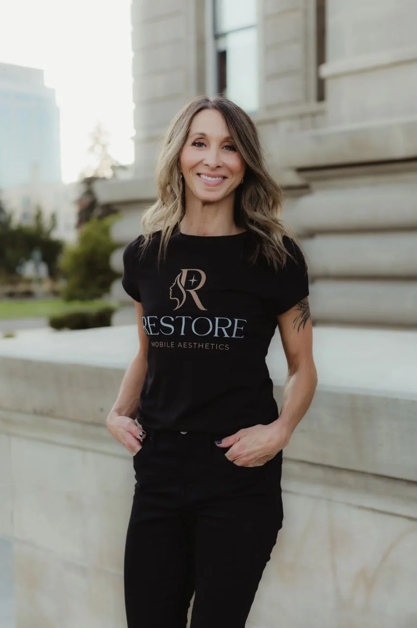 A woman with shoulder-length wavy hair smiling and wearing a black T-shirt with logo and text, standing outdoors near a building.