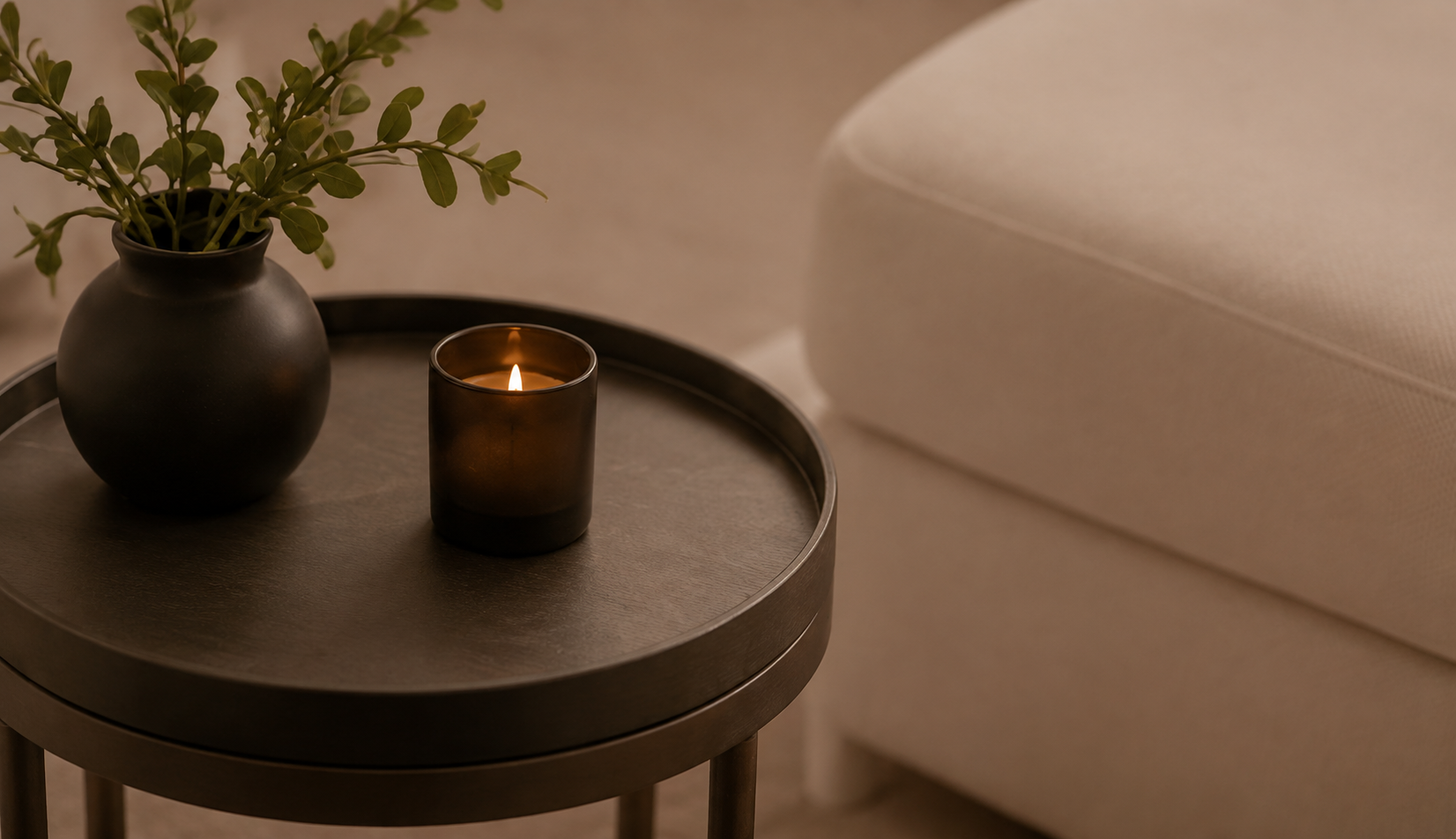 A round black side table with a black vase containing greenery, a lit brown candle in a glass holder, and a light beige sofa with textured fabric.
