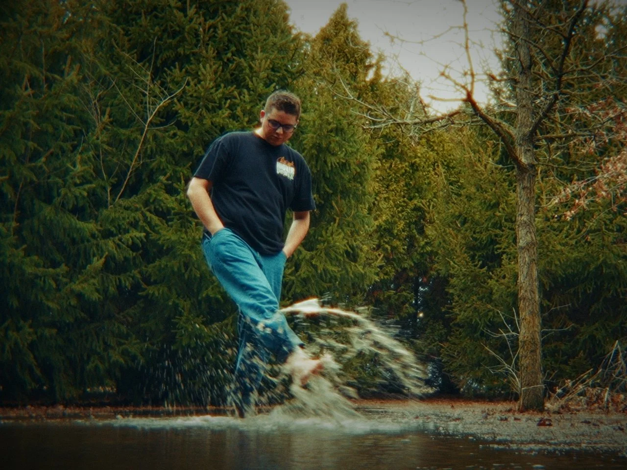 Young man walking through a puddle of water in a forested area, with trees and greenery surrounding him.
