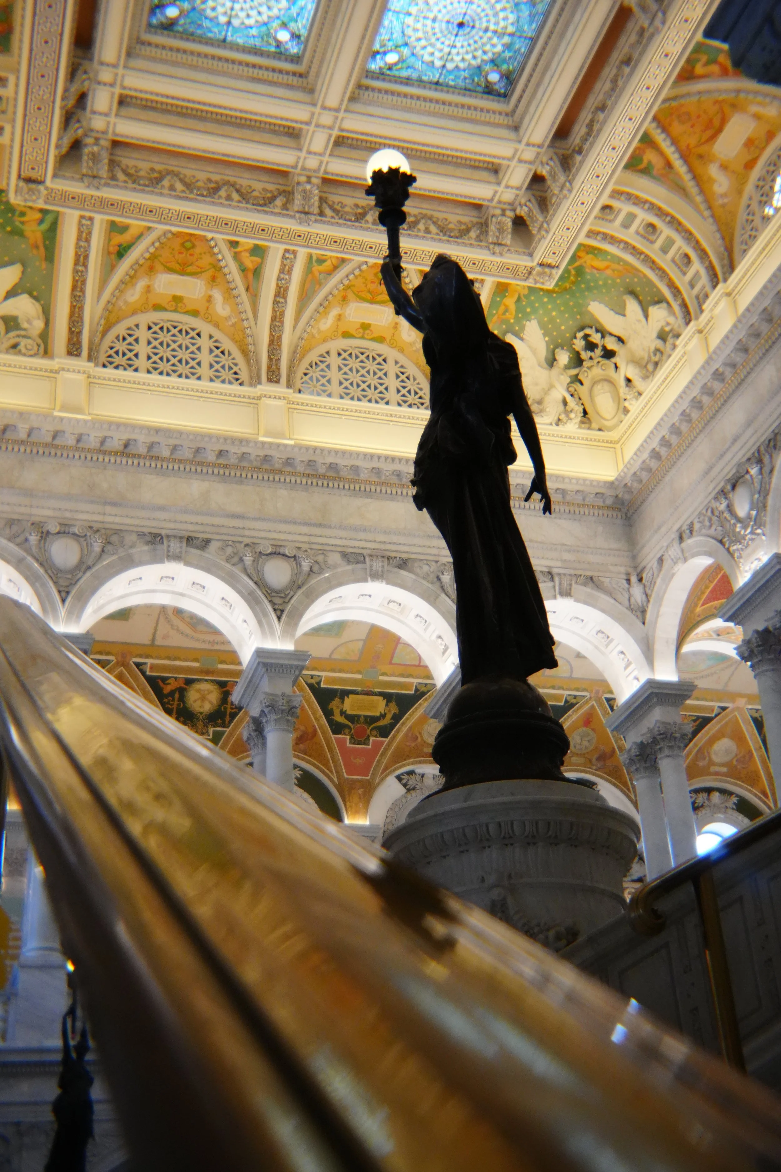 View from the bottom of a staircase looking up, showing a dark statue of a woman holding a lamp, ornate ceiling with stained glass, decorative architectural details and arches.