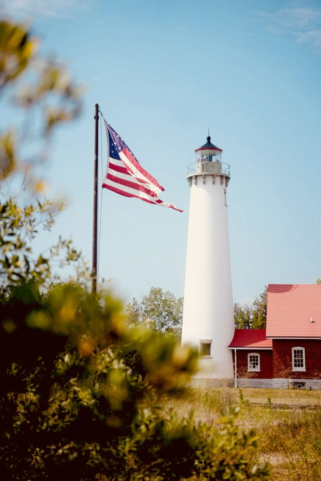 A white lighthouse with a red roof, an American flag on a pole, and some greenery in the foreground.