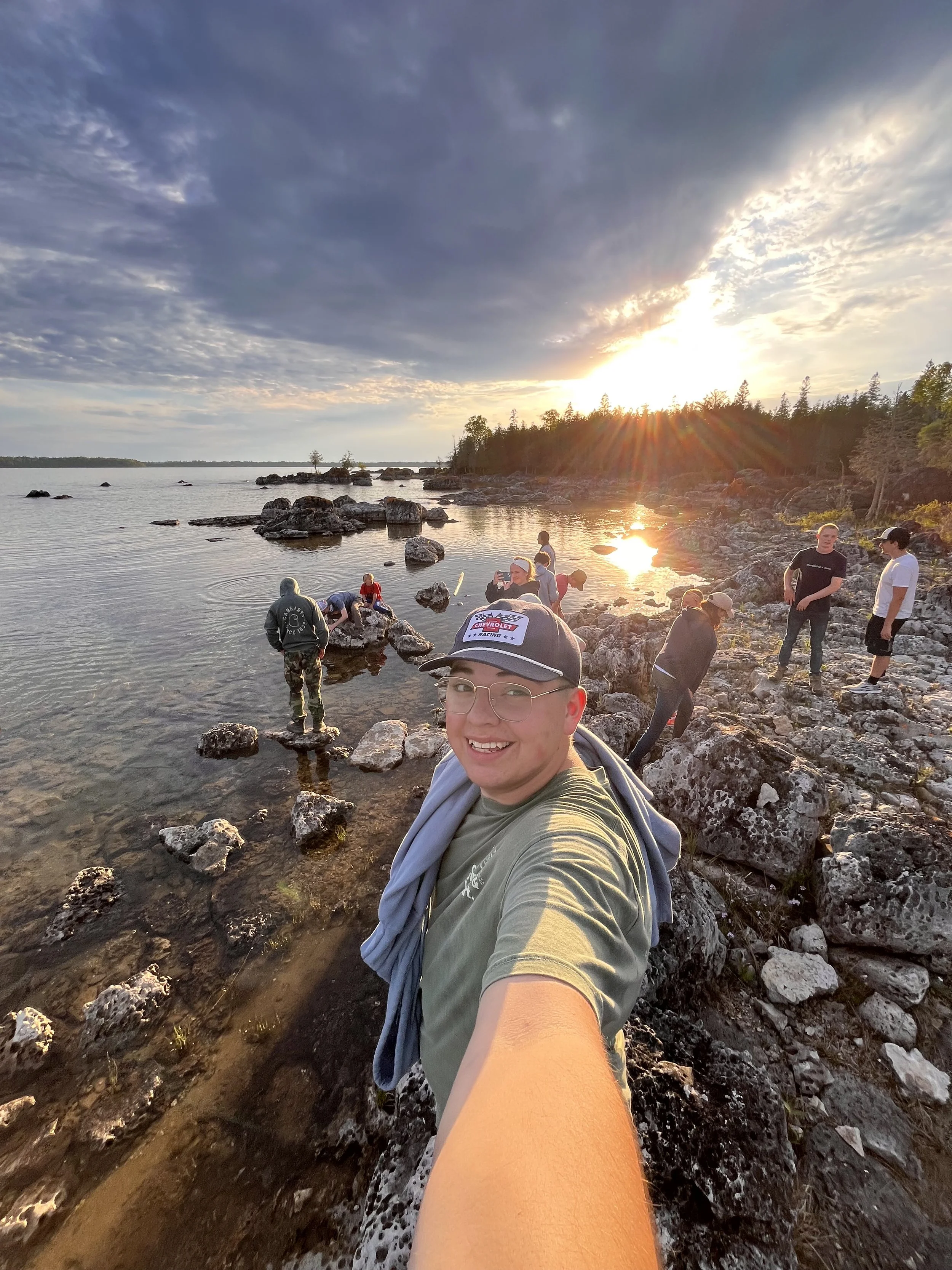Selfie of man wearing glasses and a Chevrolet racing cap, standing on rocks near a lake during sunset with other people in the background.