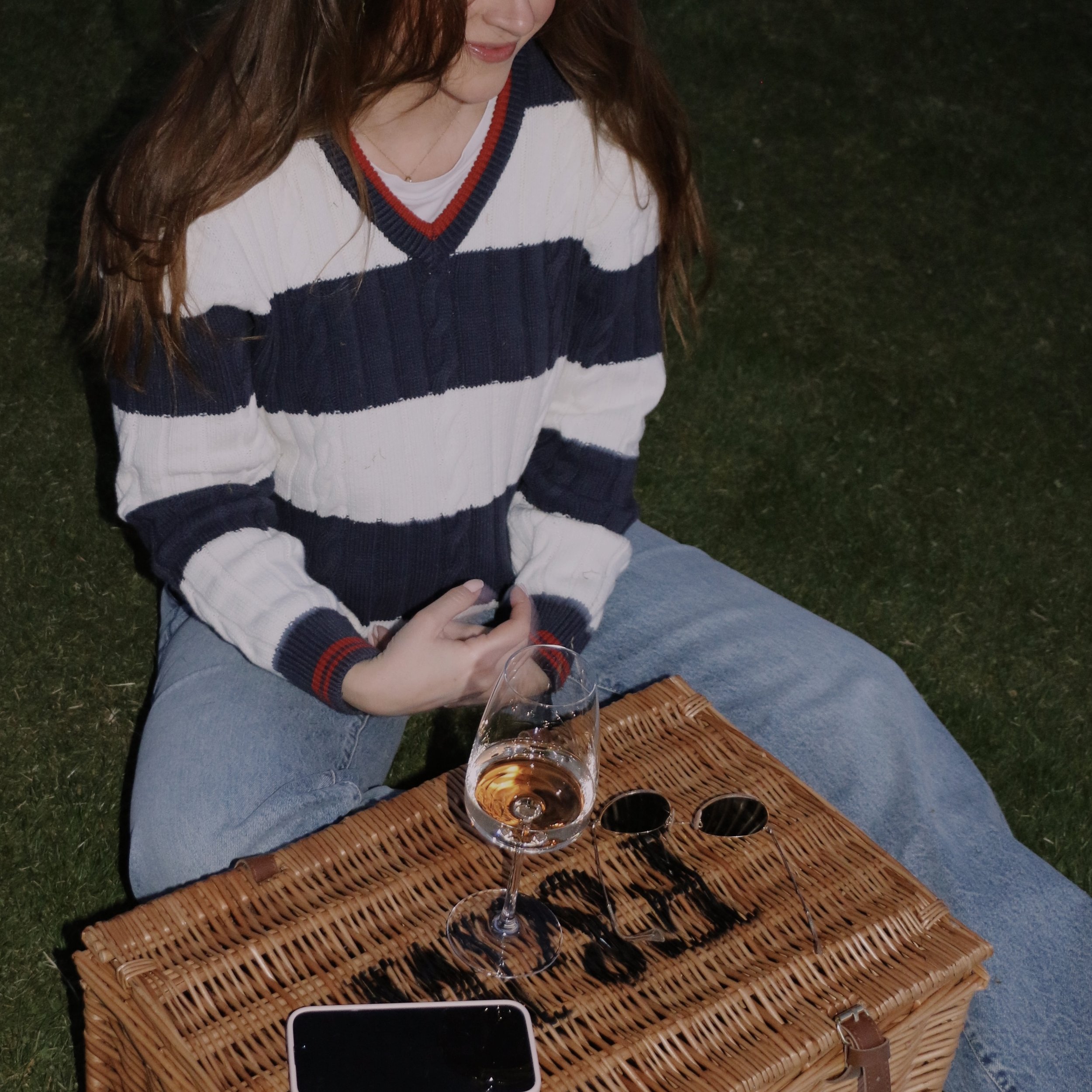 A woman sitting outdoors on a woven mat at night, holding a glass of wine with several other empty wine glasses on the mat in front of her. She is wearing a striped sweater and blue jeans, with a smartphone on the mat. She appears to be smiling or looking down, with grass visible in the background.