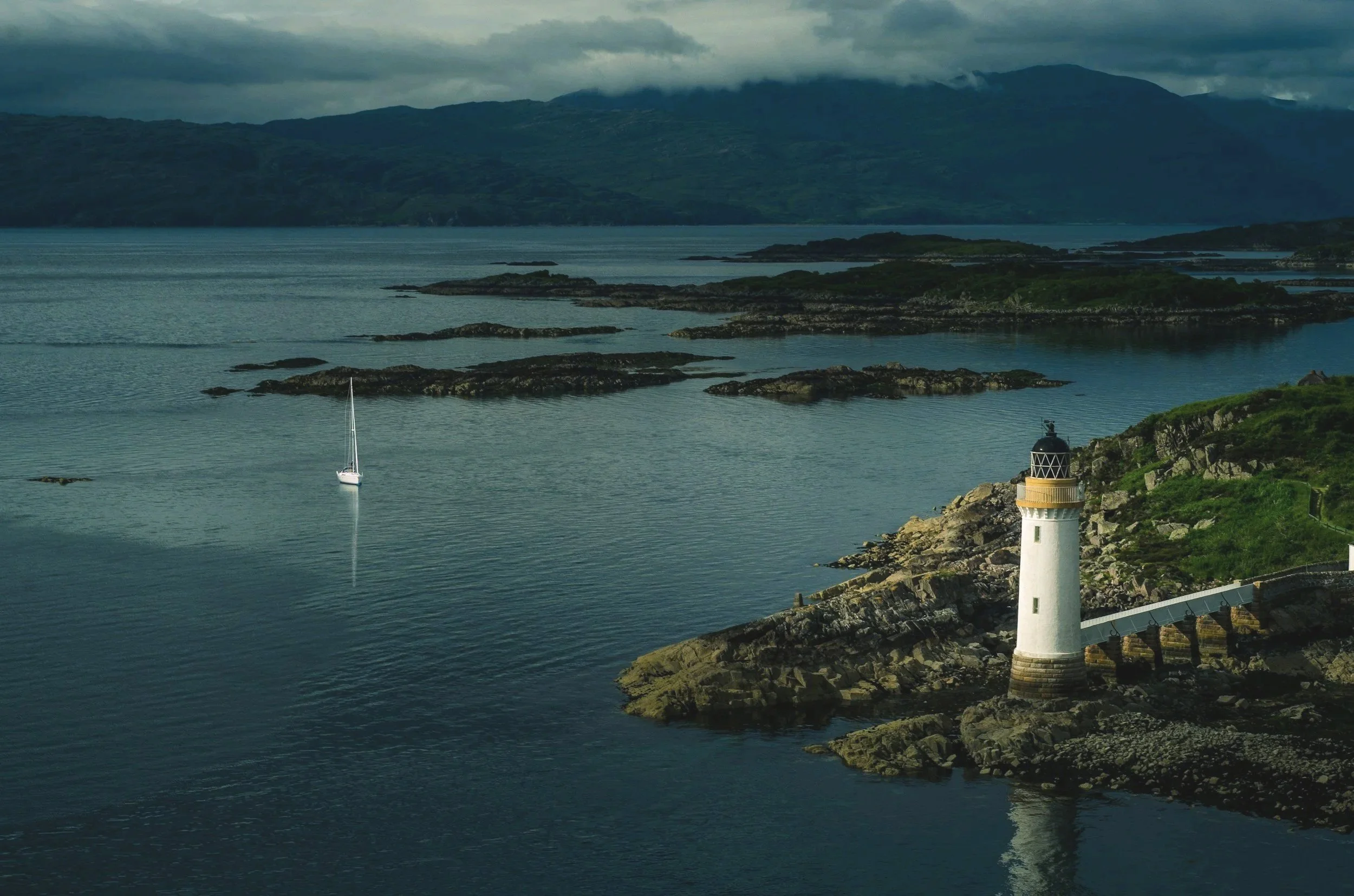 A lighthouse on a rocky coastline with a small sailboat in a body of water, and mountains in the background under cloudy skies.